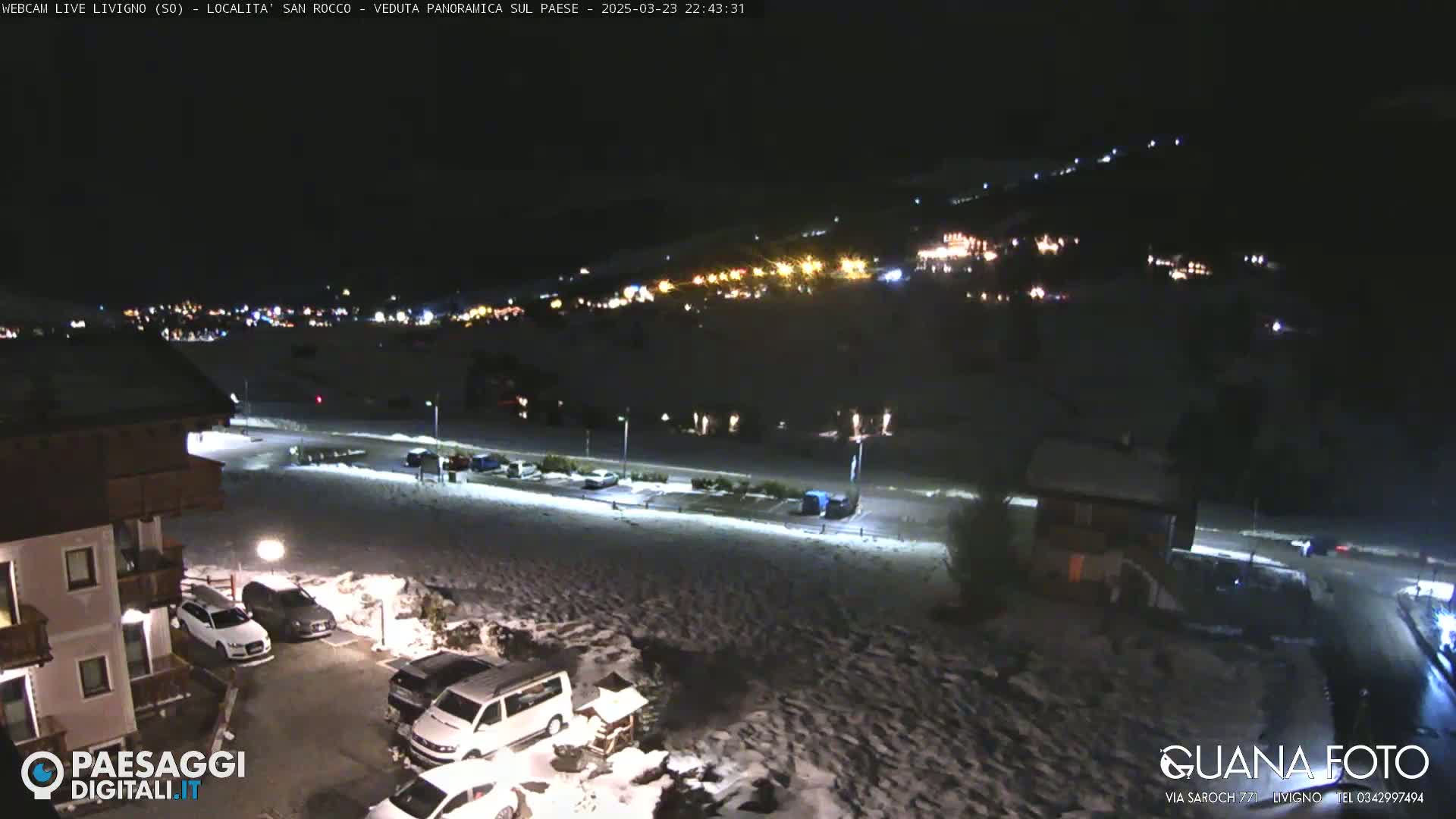 A nighttime, snow-covered parking lot and road are shown, with a few cars parked, and a hillside with illuminated buildings in the background.