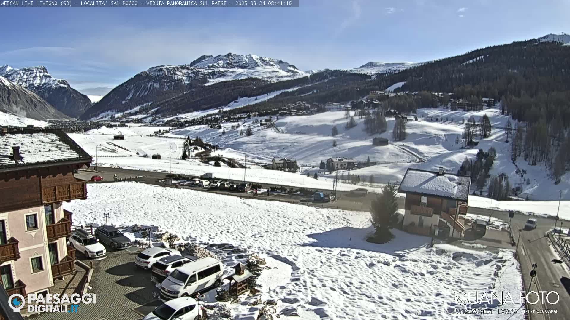 A snow-covered mountain valley scene on a sunny day, with cars parked near buildings and a road winding through the landscape.
