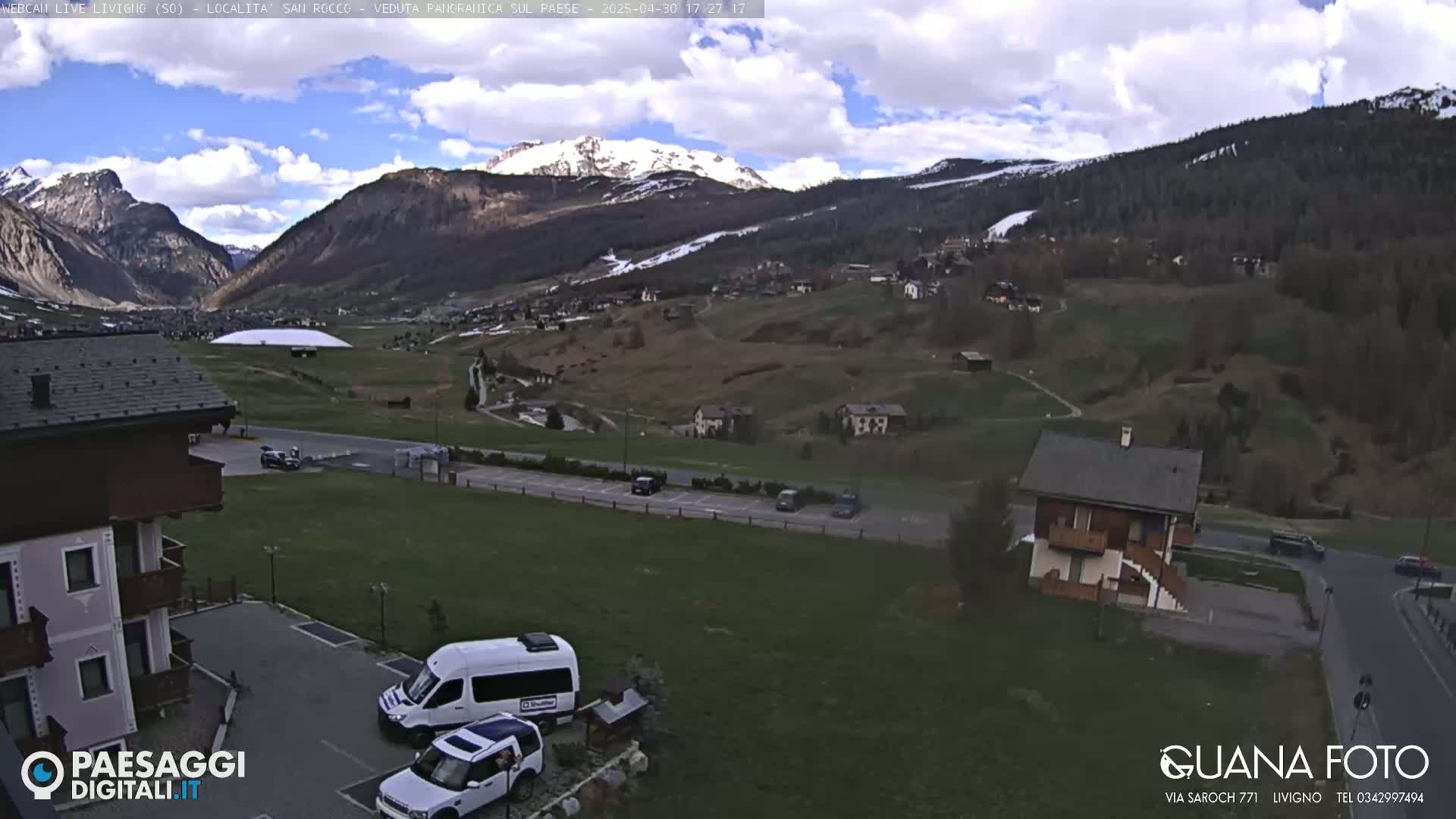 A partly cloudy day reveals a mountain valley town with snow-capped mountains in the background, viewed from a slightly elevated vantage point.