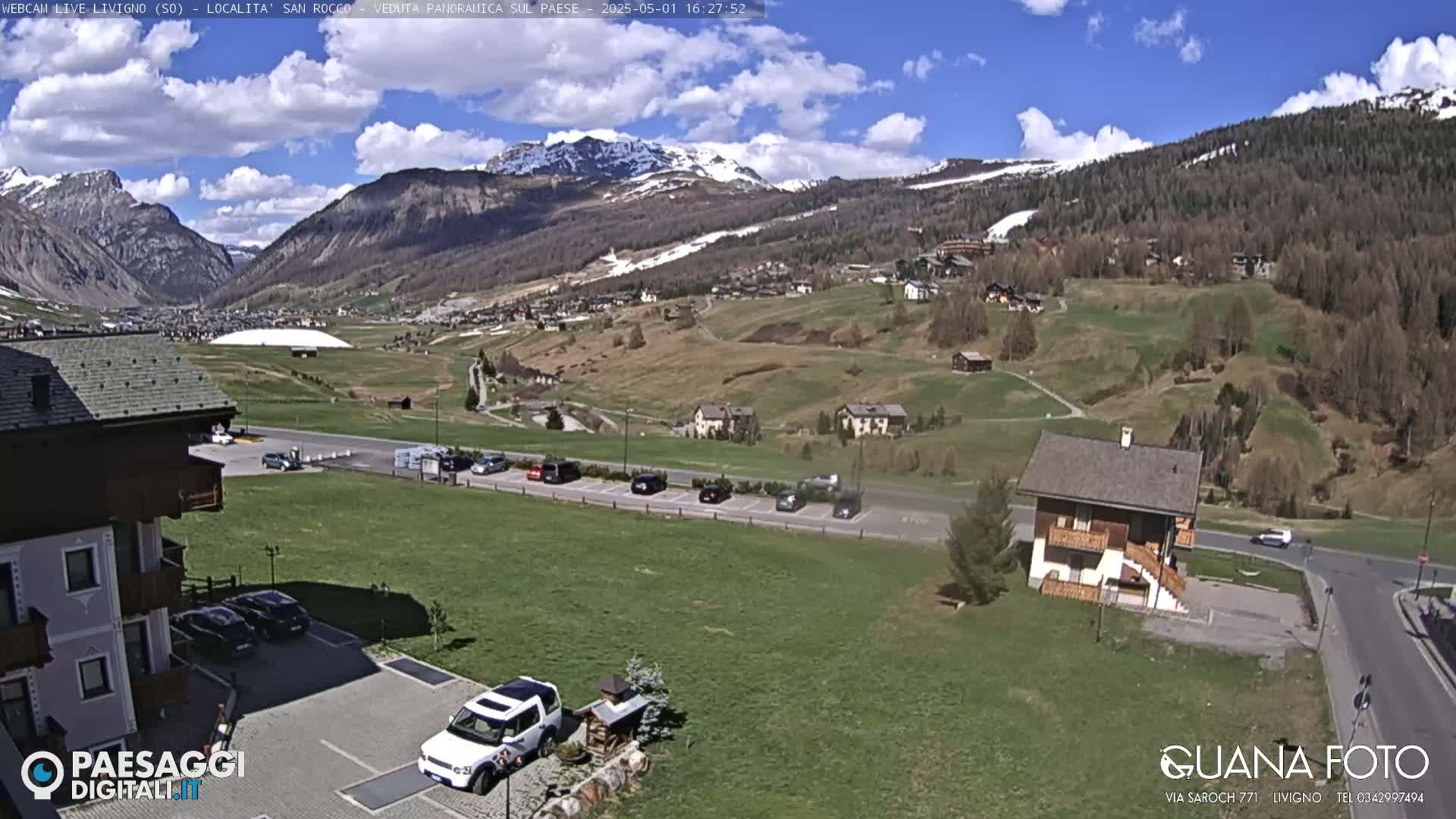 A sunny day reveals a mountainous landscape with a small village nestled in a valley, featuring a parking lot with several cars and a single-story house.