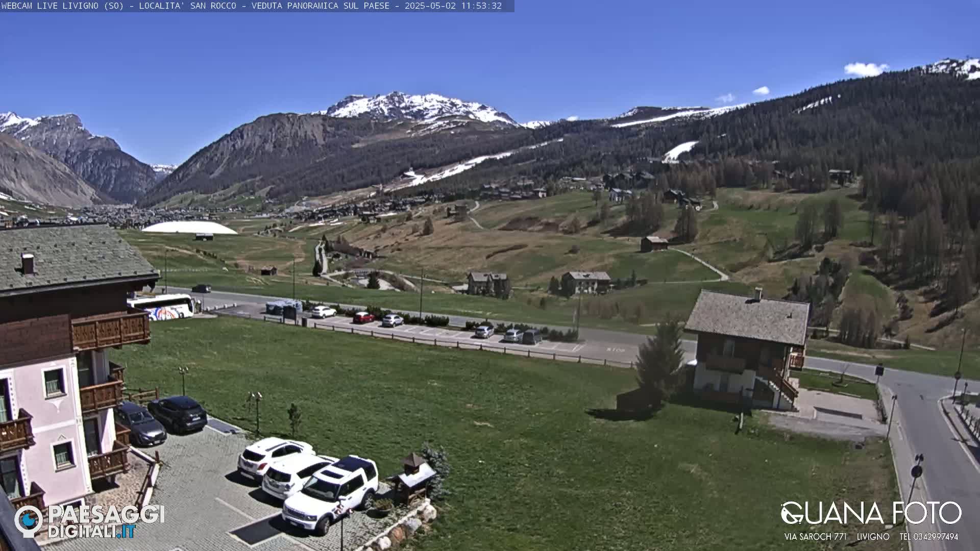 A sunny view of a mountain valley town with snow-capped mountains in the background, several cars parked near buildings, and grassy areas.