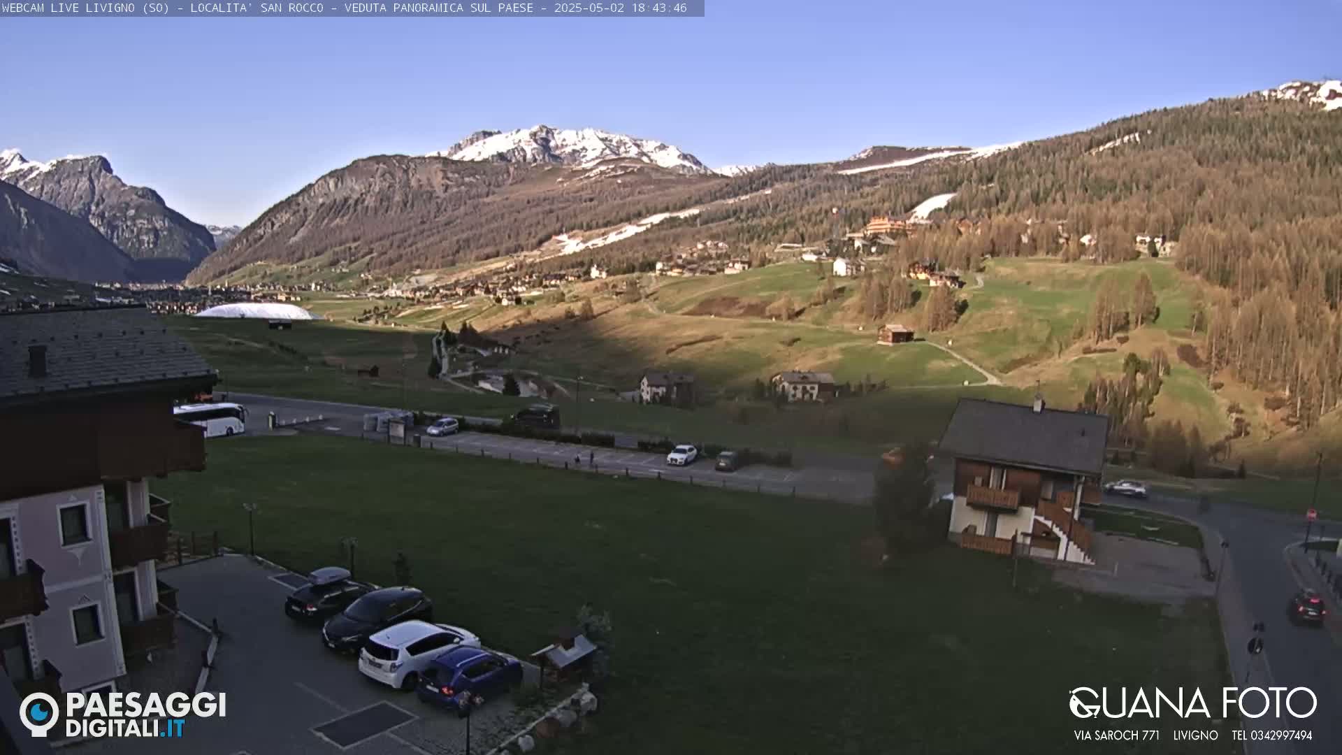 A sunny view of a mountain valley town with cars parked in a lot below, and snow-capped mountains in the background.