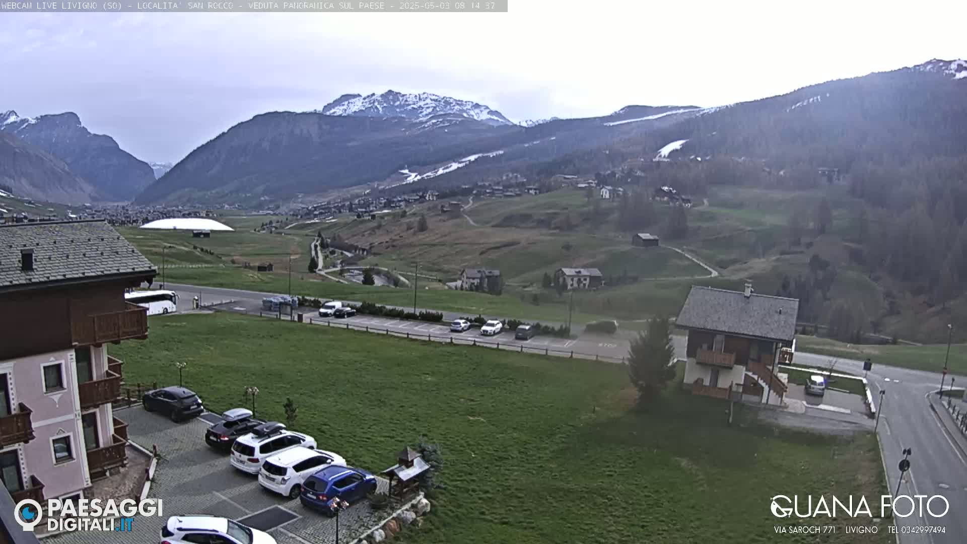 A mountain valley town with snow-capped mountains in the background, under an overcast sky, is shown with cars parked in a lot near some buildings.