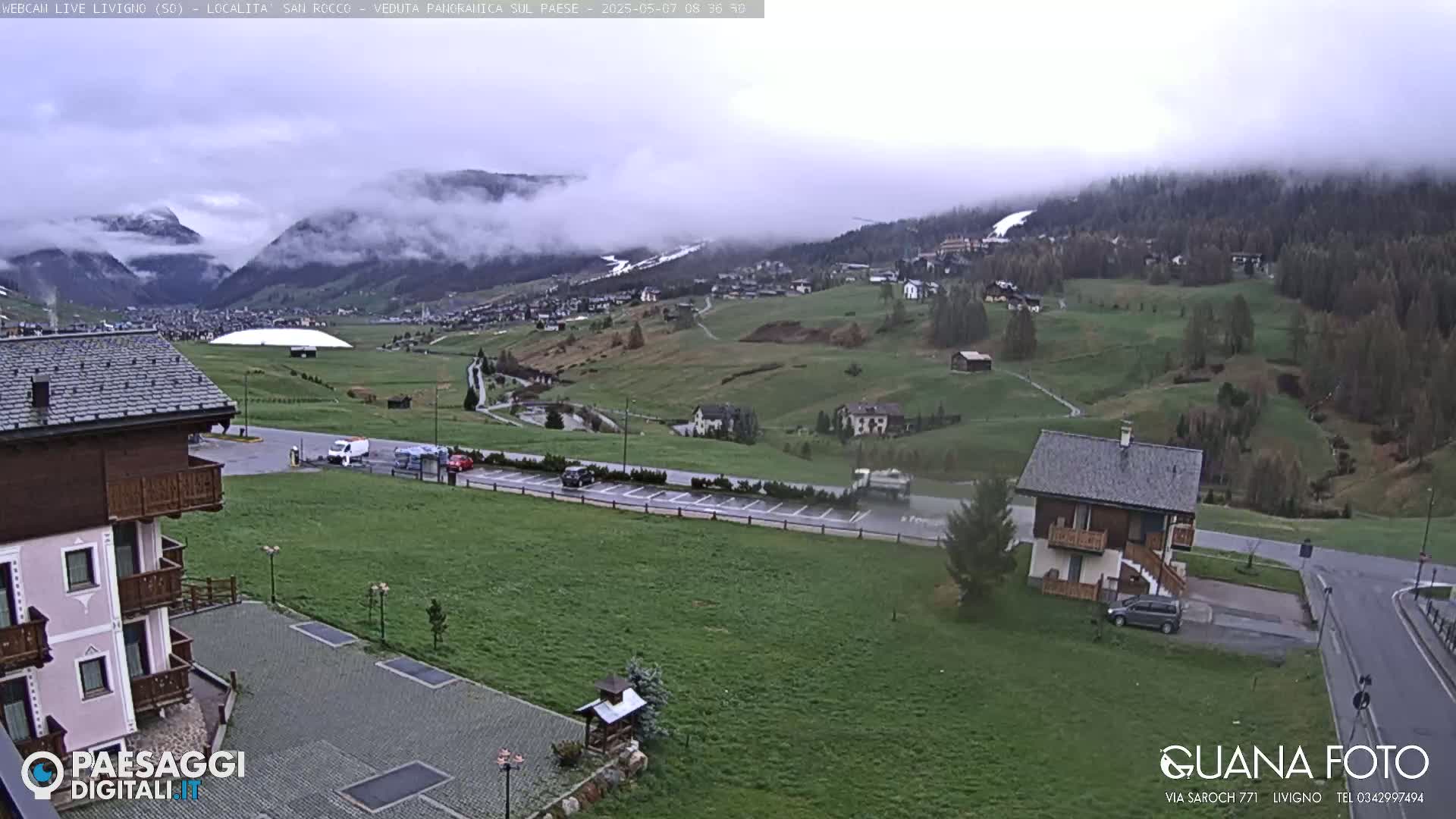 A cloudy day reveals a valley town nestled amongst green hills and mountains, with a few cars in a parking lot near a small house.