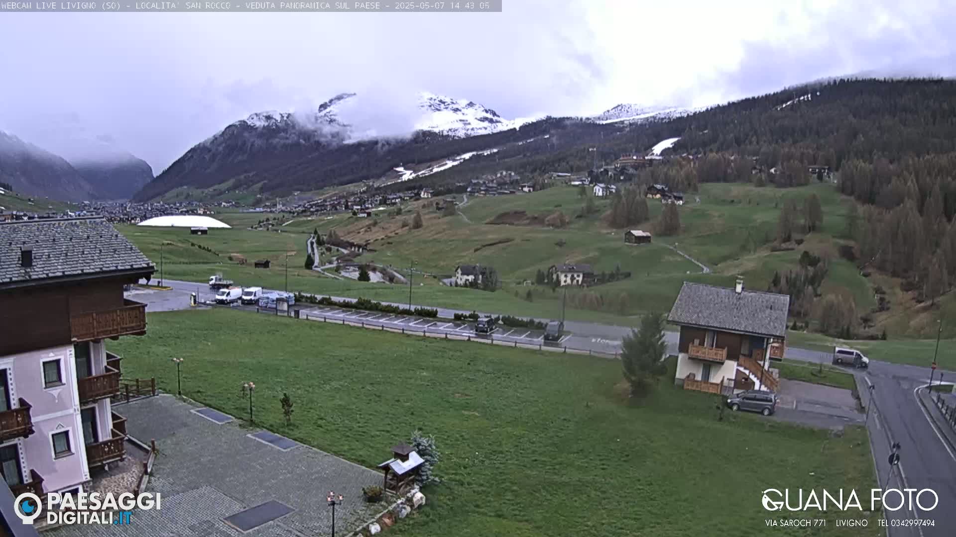 A snow-capped mountain range overlooks a valley with green fields, buildings, and a parking lot under an overcast sky.