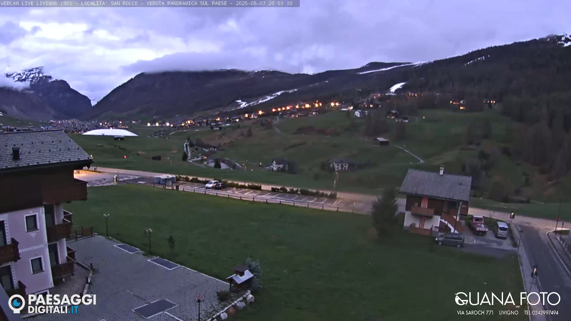 A twilight view of a mountain village with a parking lot and buildings in the foreground, under a cloudy sky.