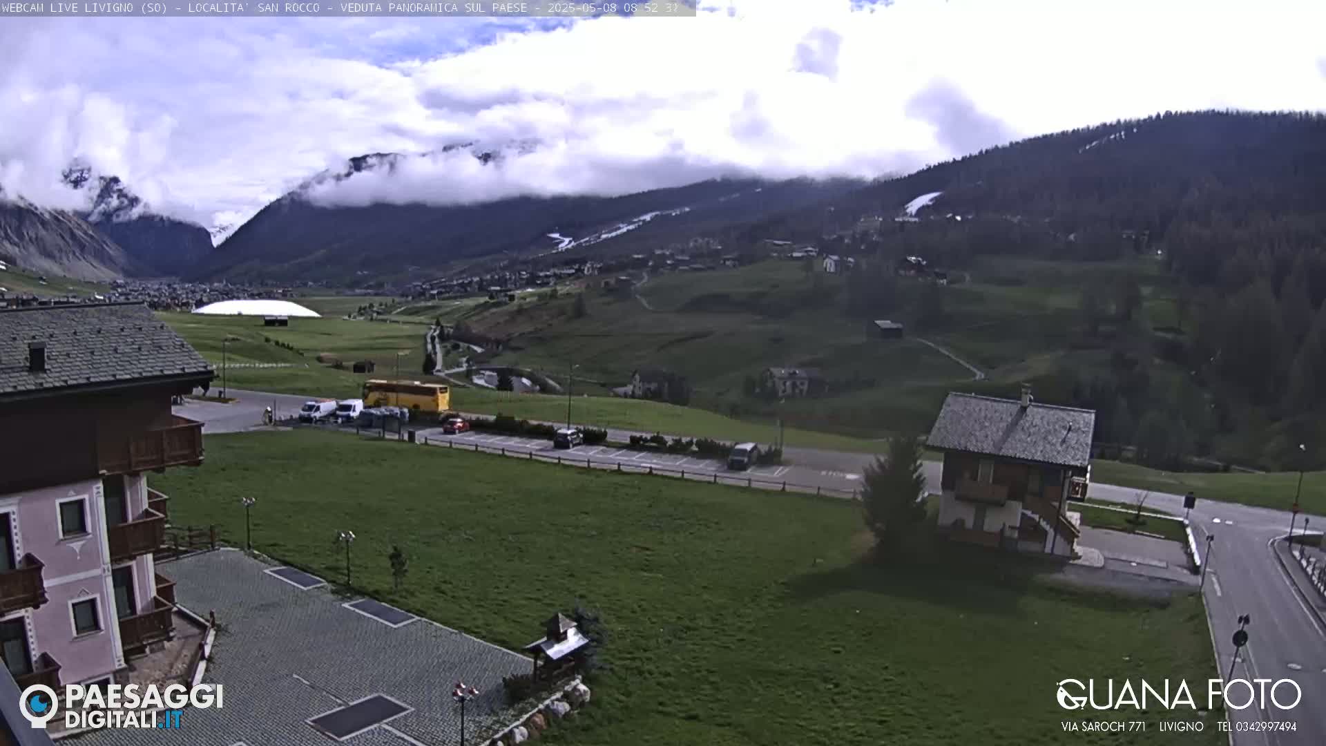 A partly cloudy day reveals a valley town nestled between mountains, with a parking lot and several buildings visible in the foreground.