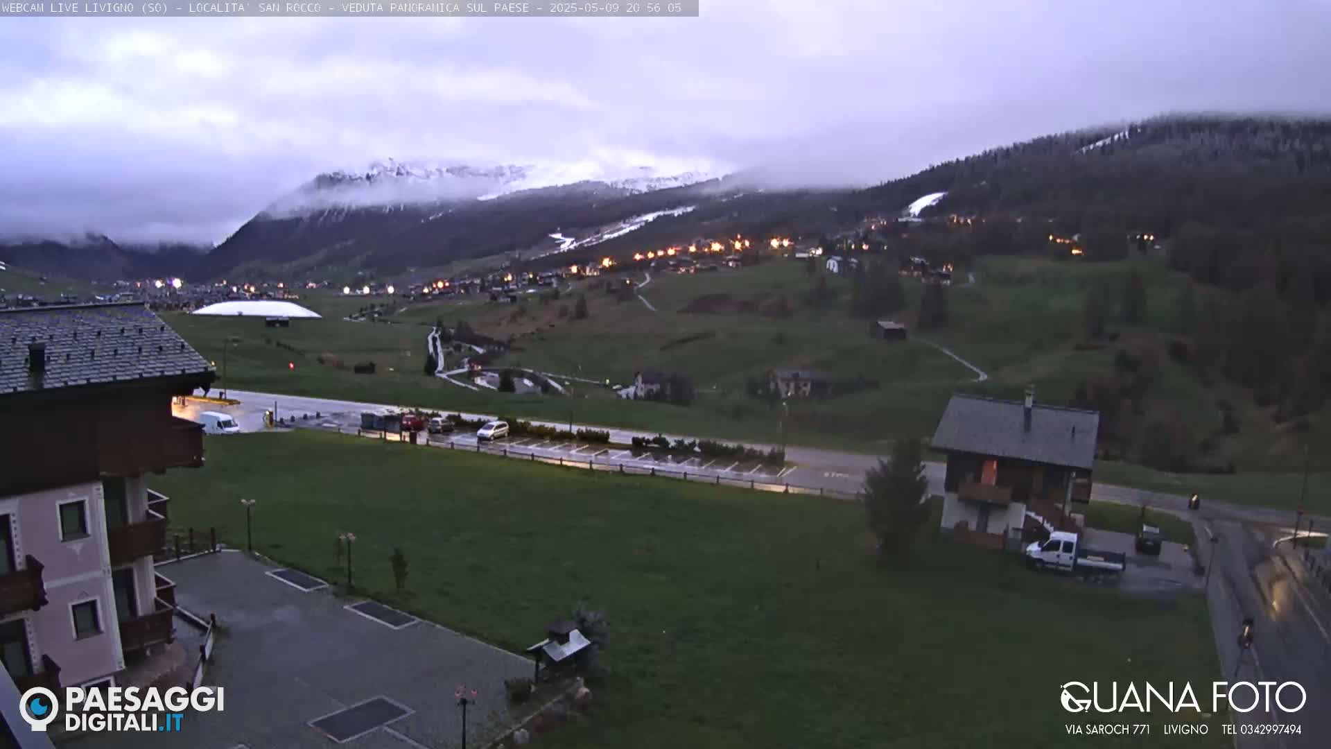 A mountain village at dusk under a cloudy sky, with snow-capped mountains in the background and a few cars in a parking lot.