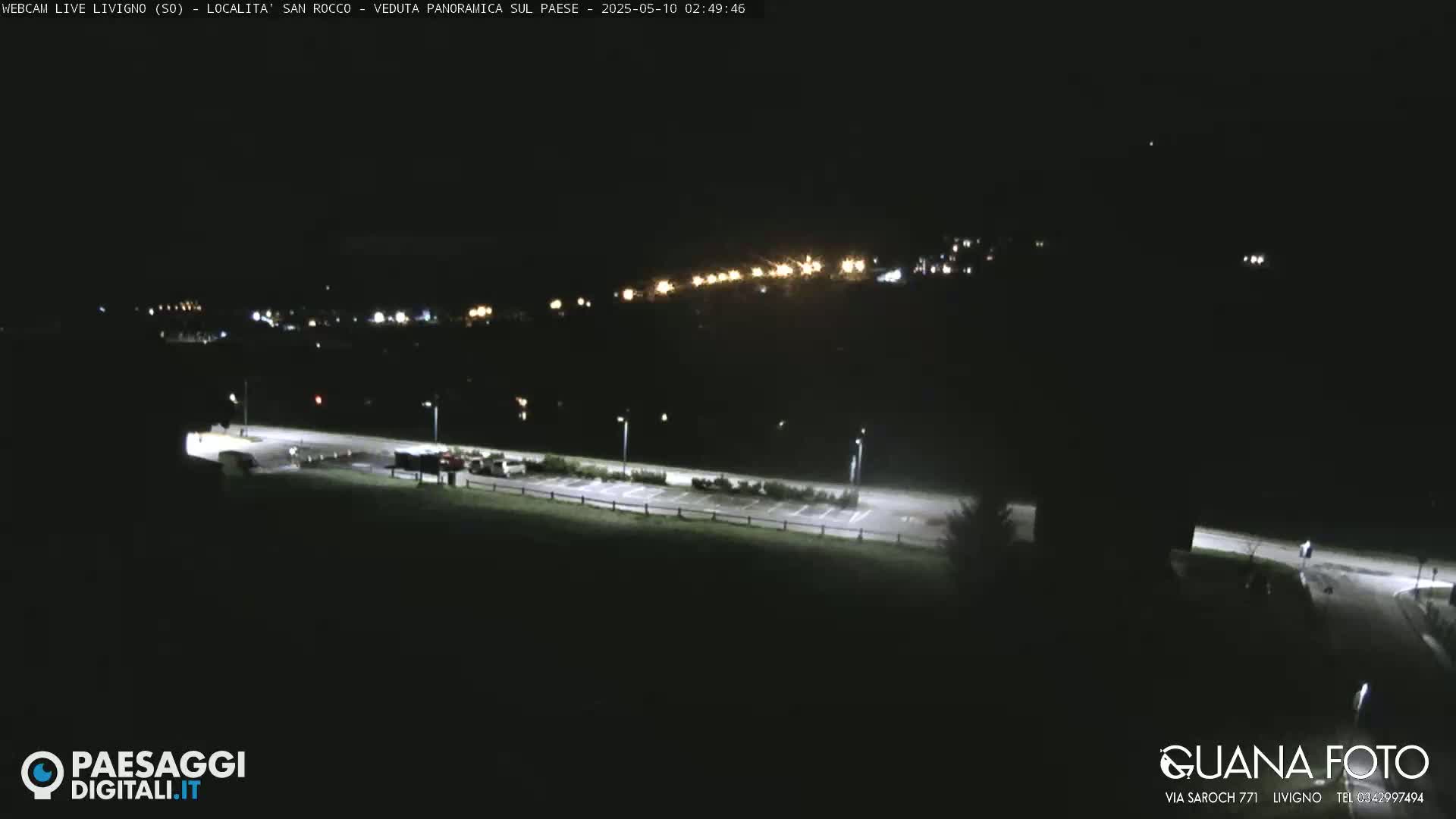 A nighttime, long shot of a mostly empty parking lot with a town's lights visible in the distance.