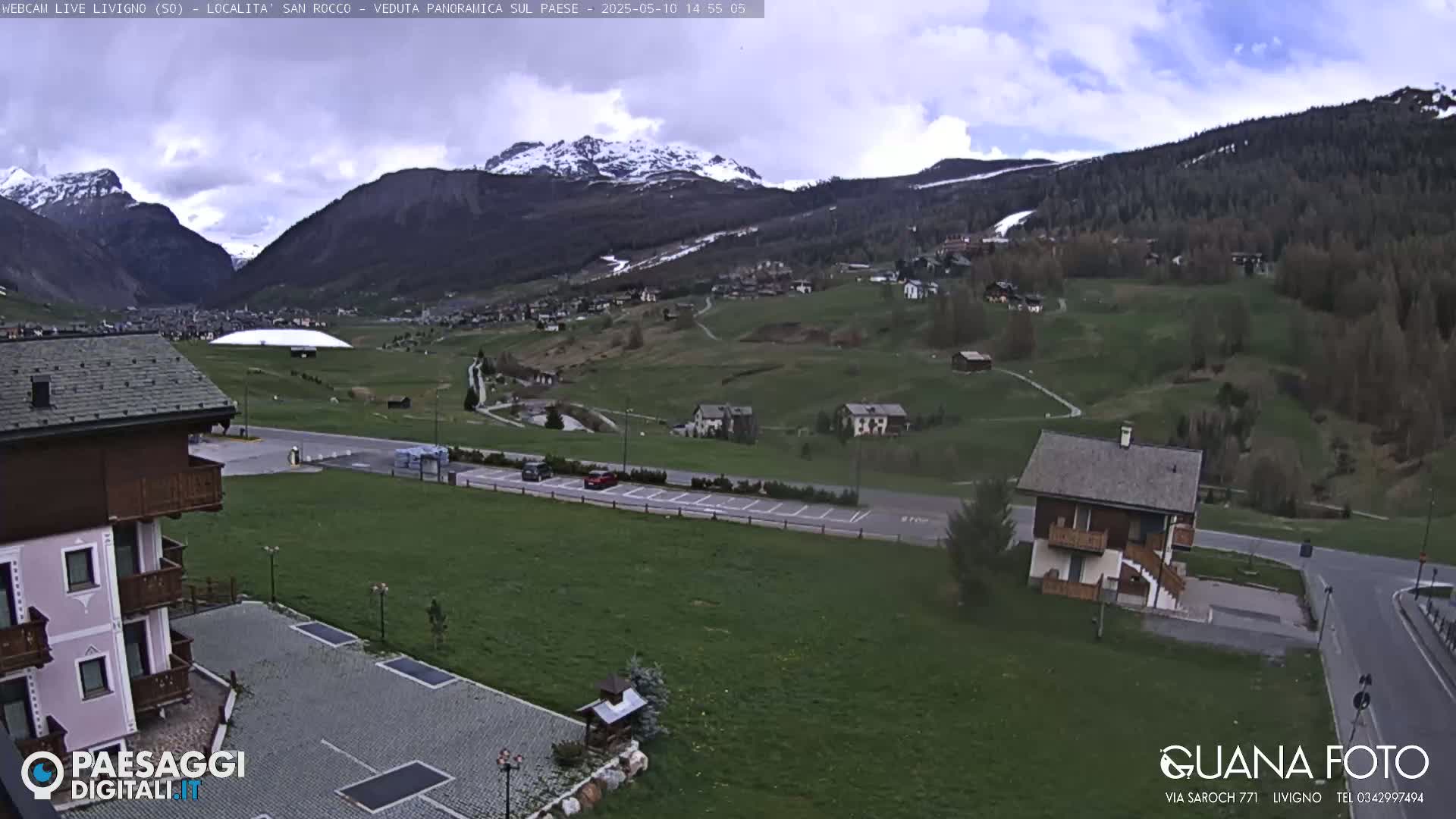A mountain valley village with snow-capped mountains in the background under a partly cloudy sky.