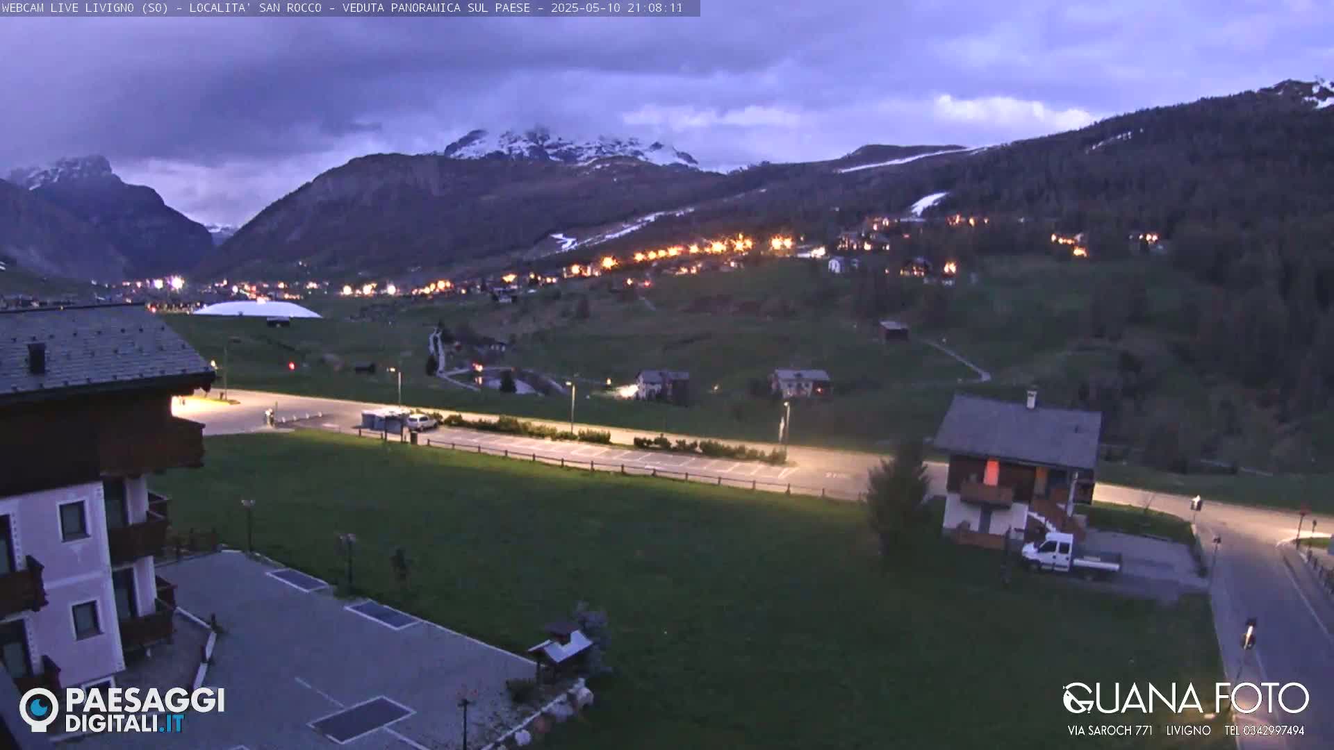 A twilight view shows a small town nestled in a valley between snow-capped mountains.