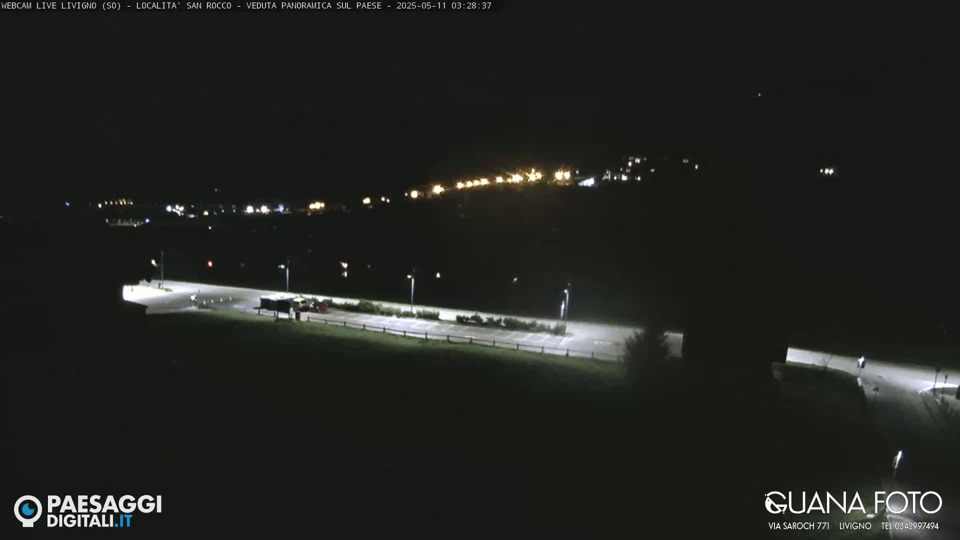 A nighttime, panoramic view of a mostly empty parking lot with a hilltop town's lights visible in the distance.