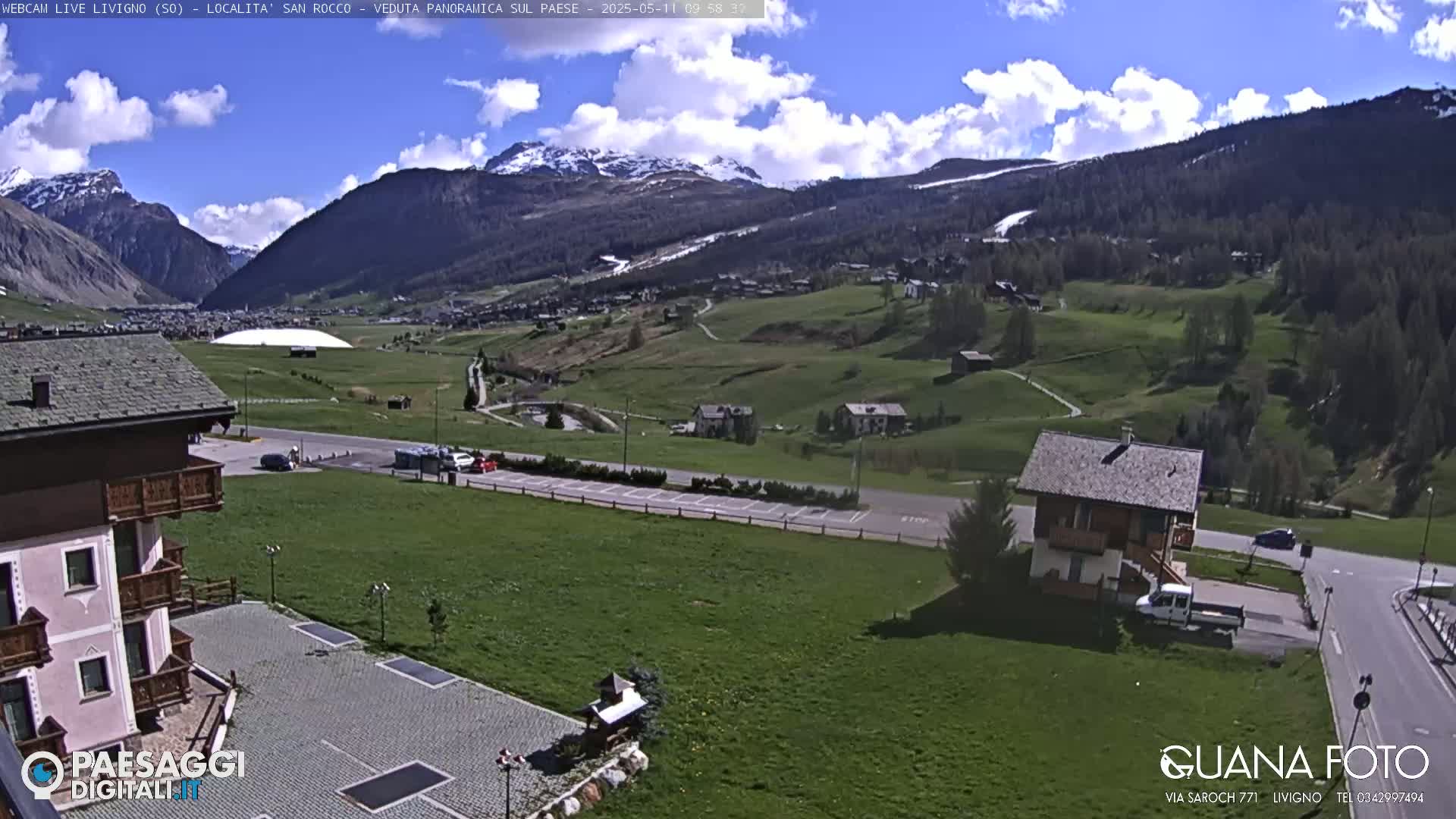 A partly cloudy day reveals a valley town nestled amongst snow-capped mountains and green hills.
