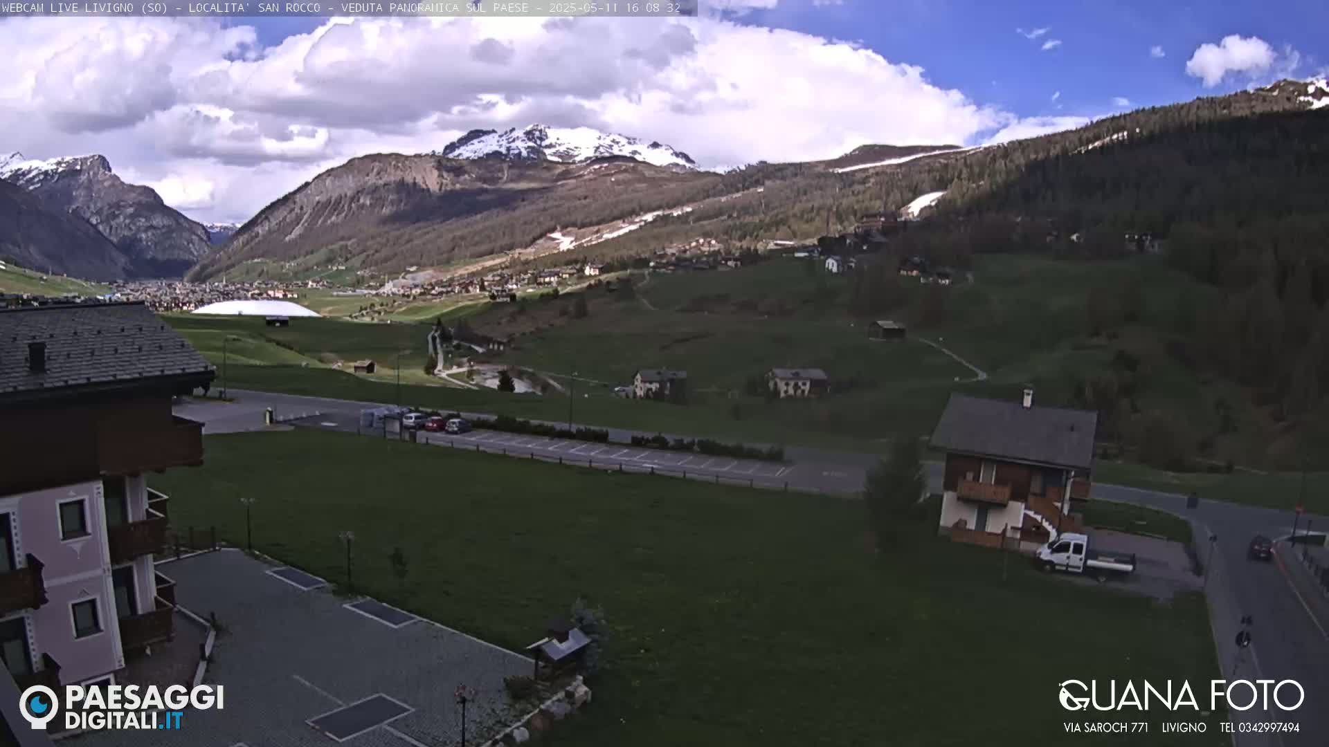 A partly cloudy day reveals a mountain valley village with a large, domed building in the distance, and a small house and parking lot in the foreground.