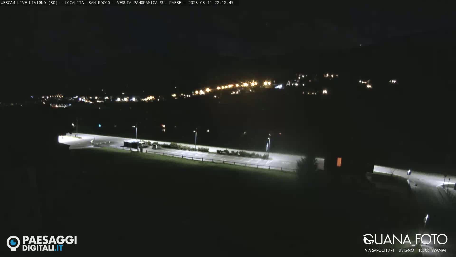 A nighttime, long shot of a mostly empty parking lot with a village's lights visible on a hillside in the distance.
