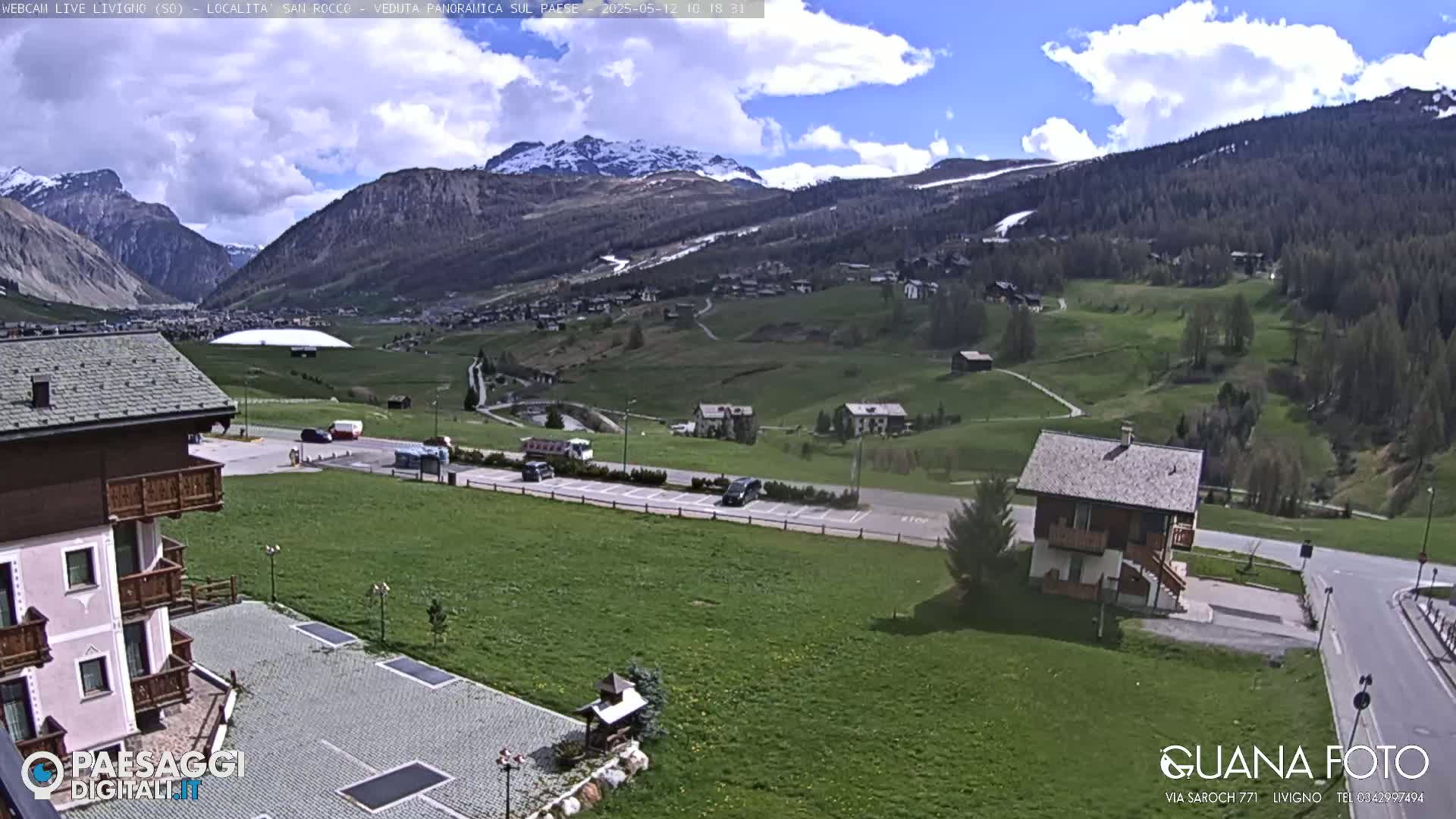 A partly cloudy day reveals a mountain valley town with green fields, a parking lot, and several buildings, nestled among snow-capped mountains.