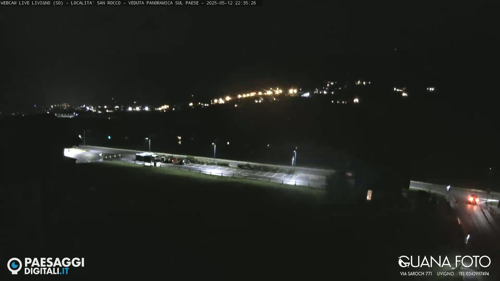 A nighttime view of a hilltop town illuminated by lights, a road, and a mostly empty parking lot below.