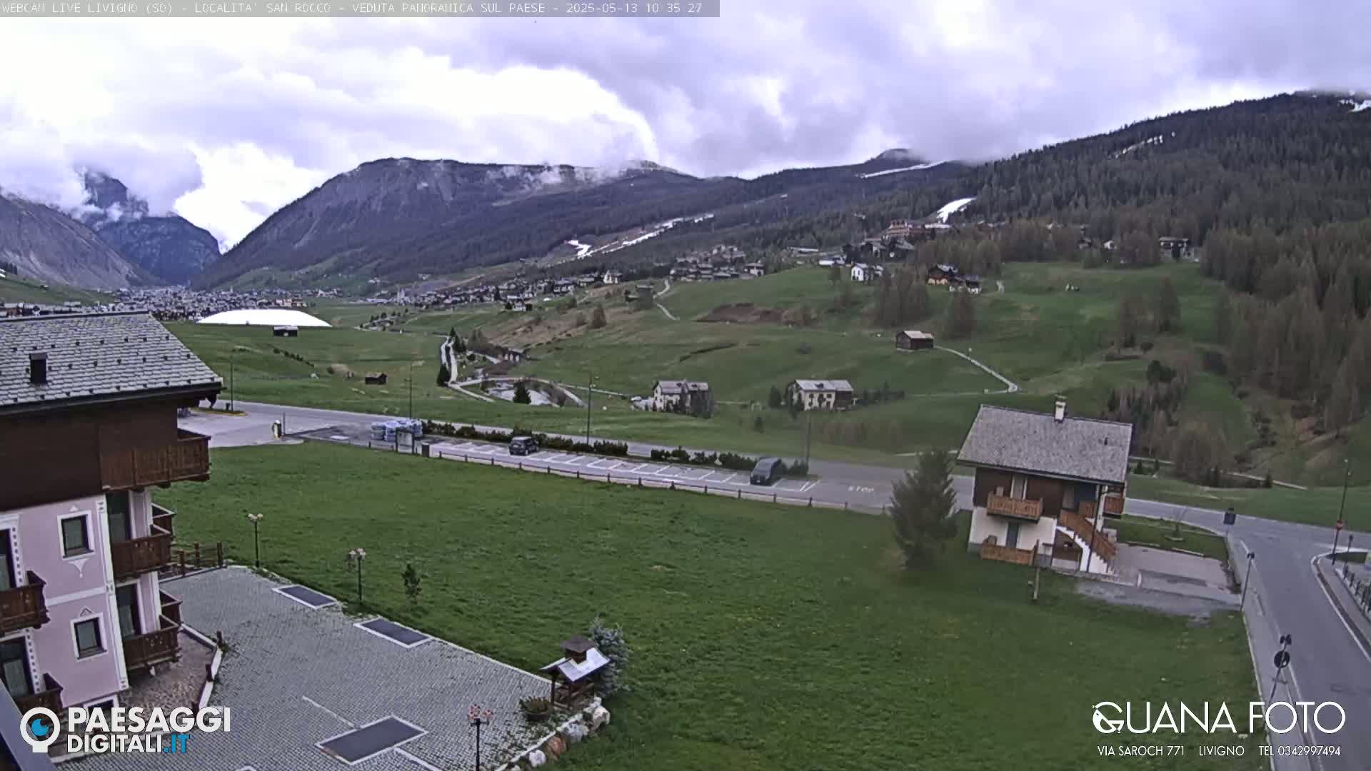 A cloudy day reveals a mountain village nestled in a valley, with green fields and a few buildings visible, and some cars parked on roads nearby.