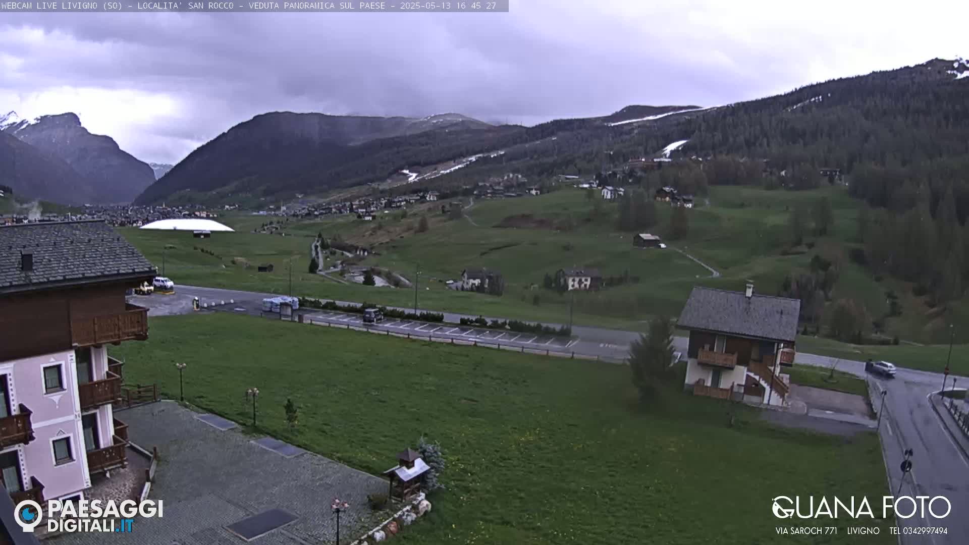 A mountain valley town is visible under an overcast sky, with green fields and buildings scattered throughout, and snow-capped mountains in the background.