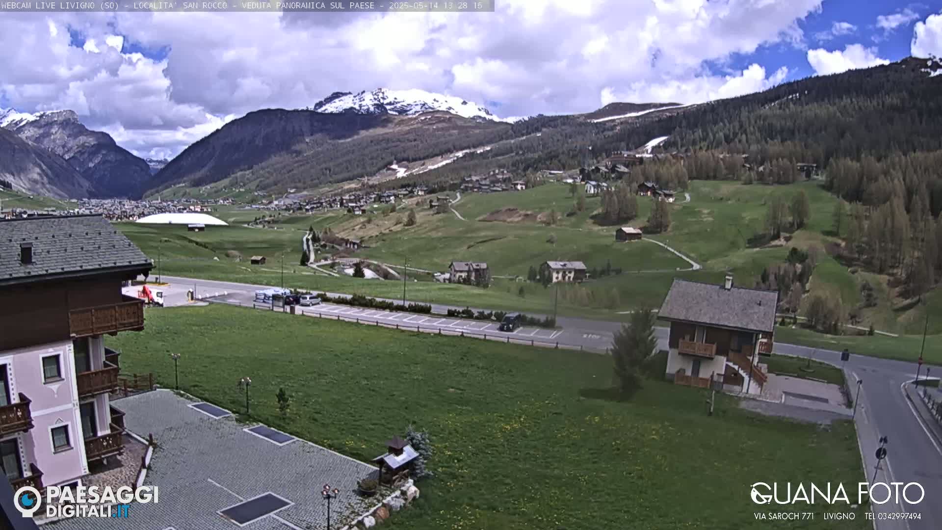 A partly cloudy day reveals a mountain valley village with green hillsides, a small house, and a parking lot, with snow-capped mountains in the background.