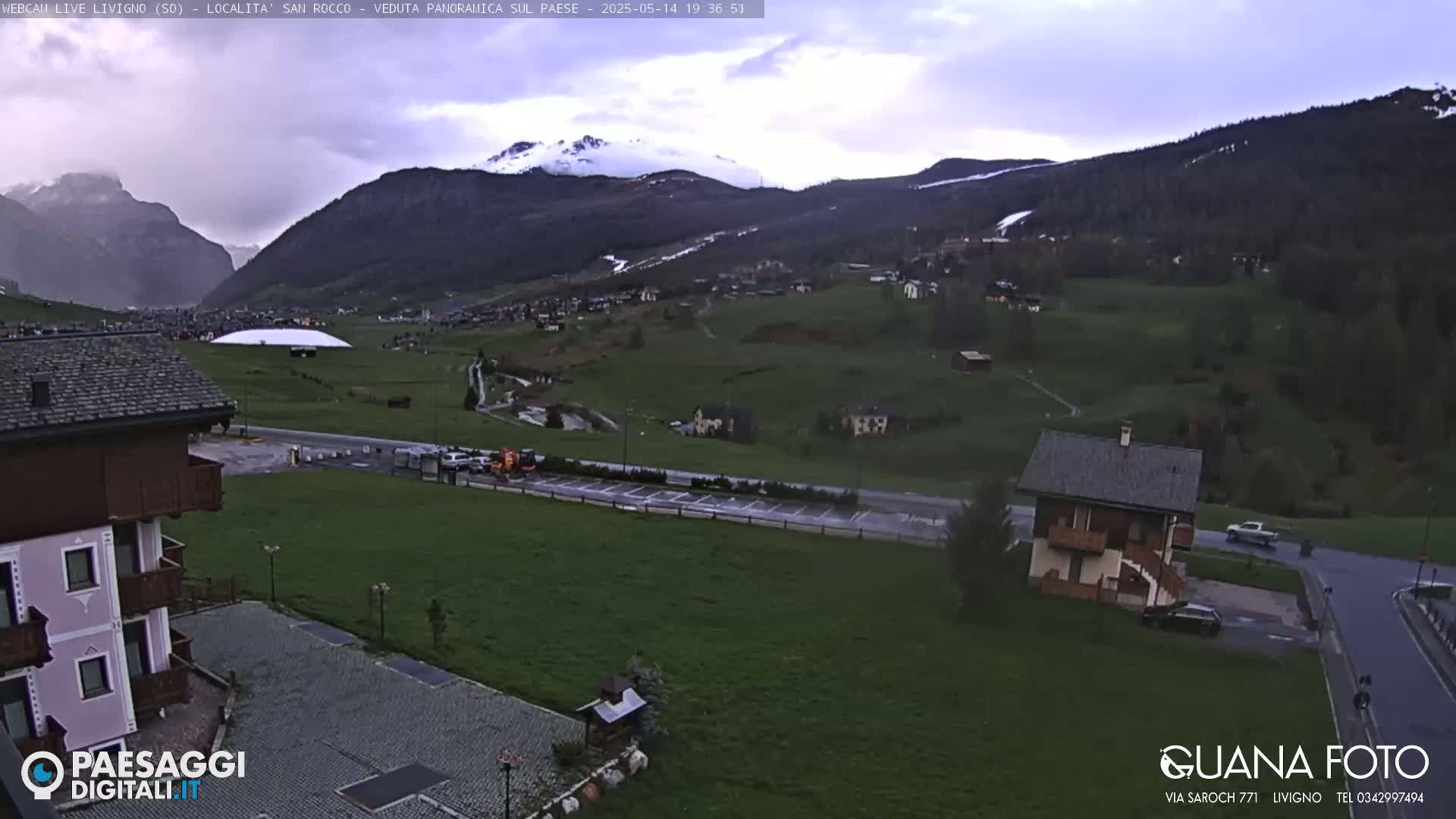 A mountain village under an overcast sky, with snow visible on distant peaks, is viewed from an elevated position, showing a parking lot, buildings, and green fields.