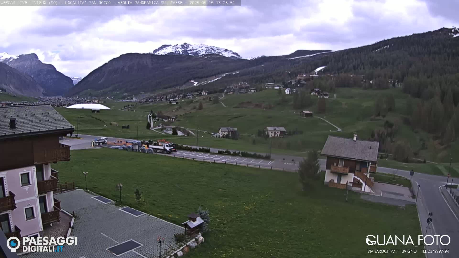 A mostly cloudy day reveals a village nestled in a valley between snow-capped mountains, with a large building resembling a dome visible in the middle ground and a small house in the foreground.