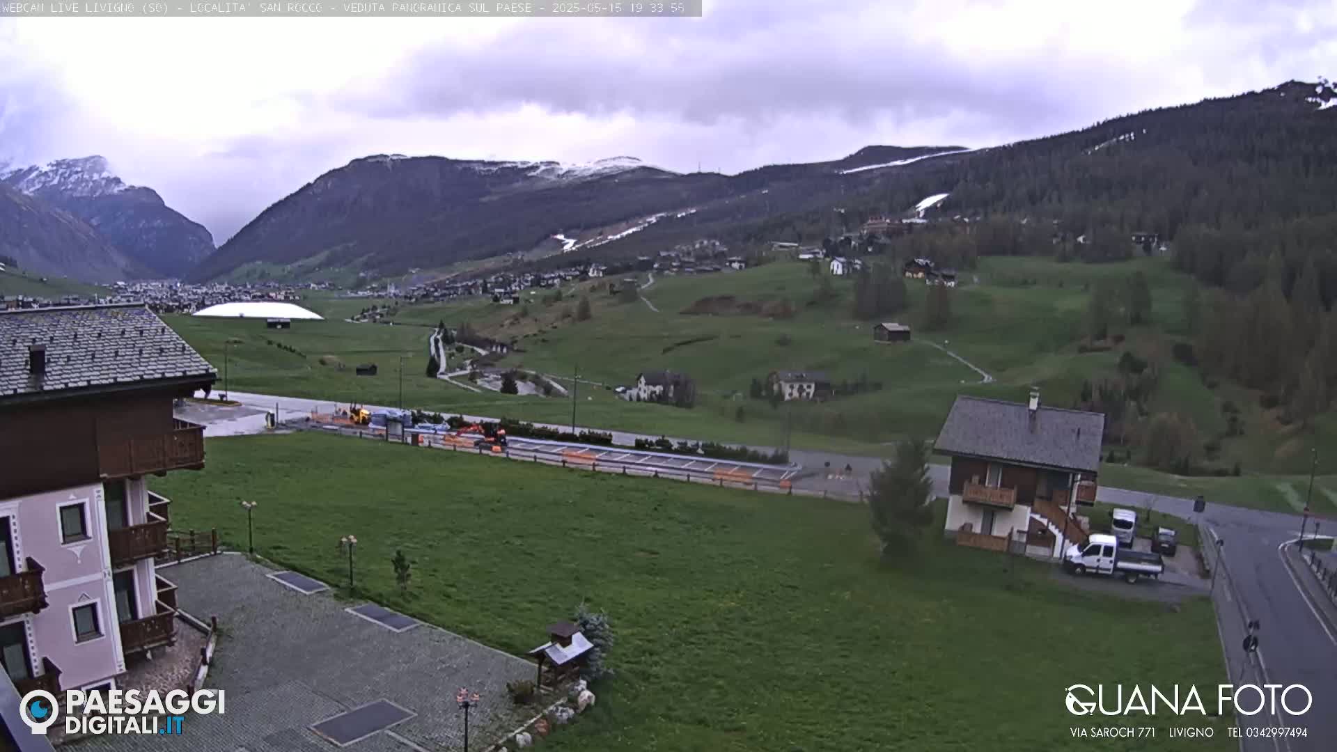 A cloudy day reveals a valley town nestled among green hills and snow-capped mountains, with a large white structure in the distance.