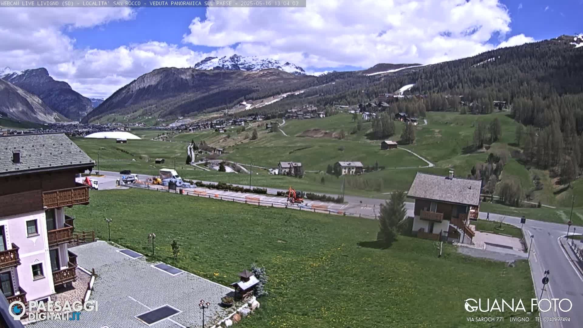 A partly cloudy day shows a wide, grassy valley with a few buildings, a parking lot, and snow-capped mountains in the background.