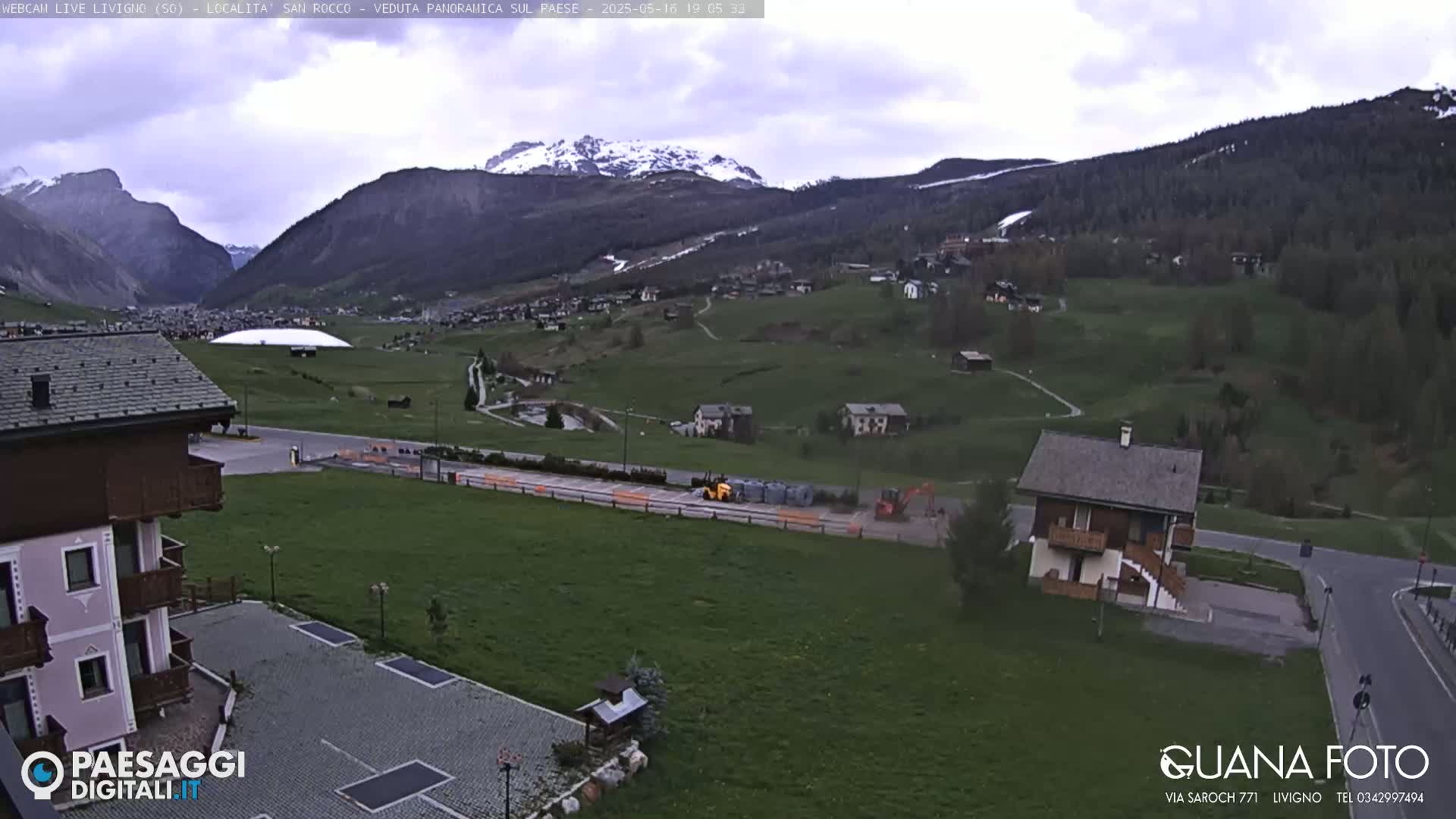 A partially cloudy day reveals a mountain valley town nestled among green hills and snow-capped mountains.