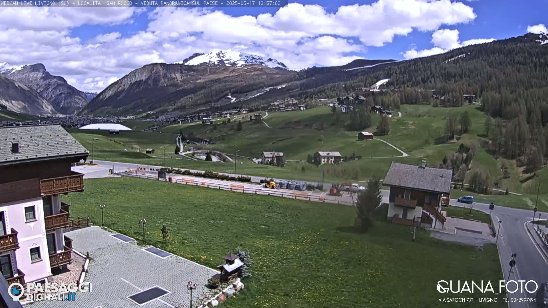 A partly cloudy day reveals a valley town nestled among green hills and snow-capped mountains, with construction equipment visible in a parking lot.