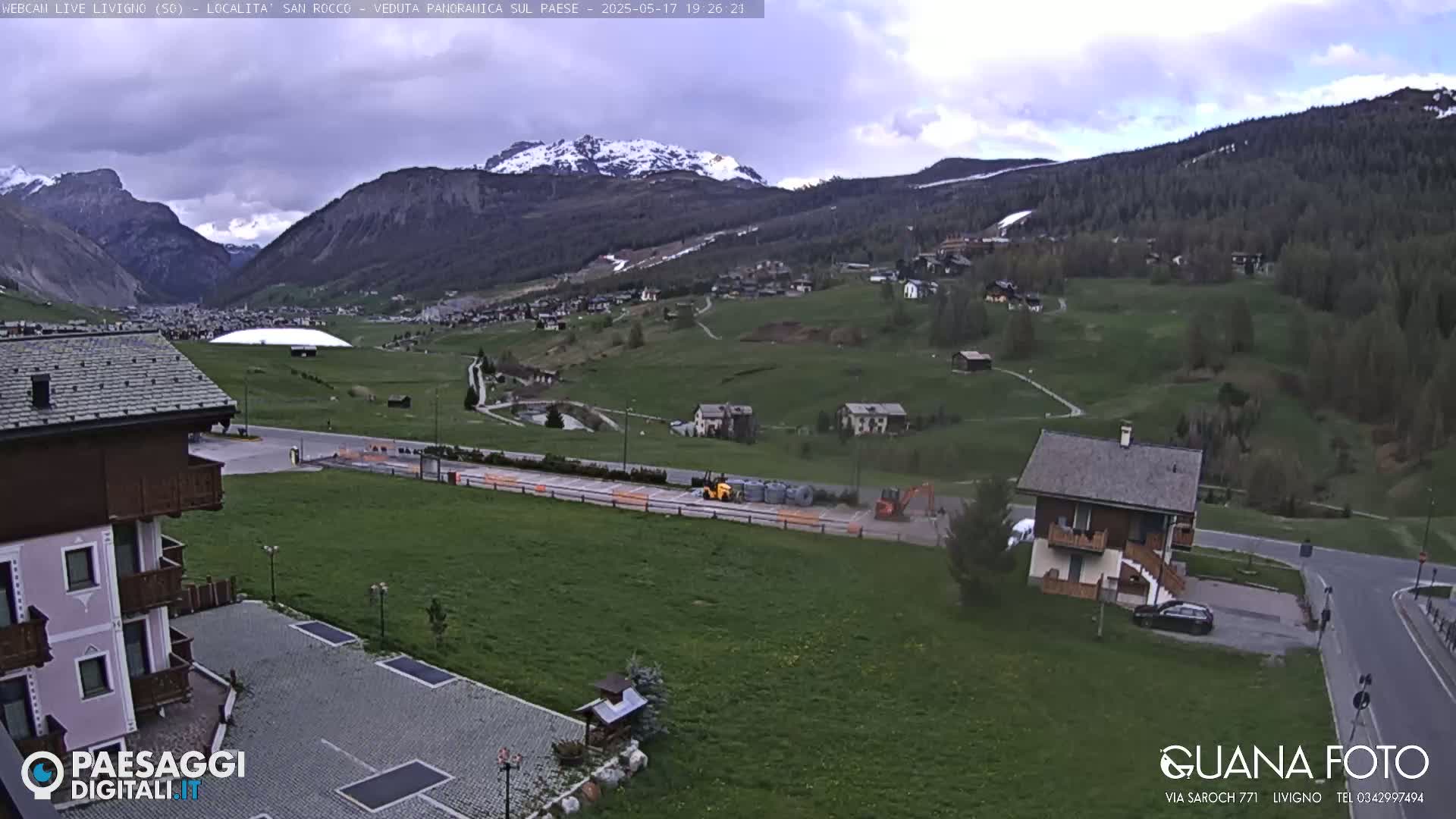 A mountain valley town with green fields, a few buildings, and some construction equipment under an overcast sky.