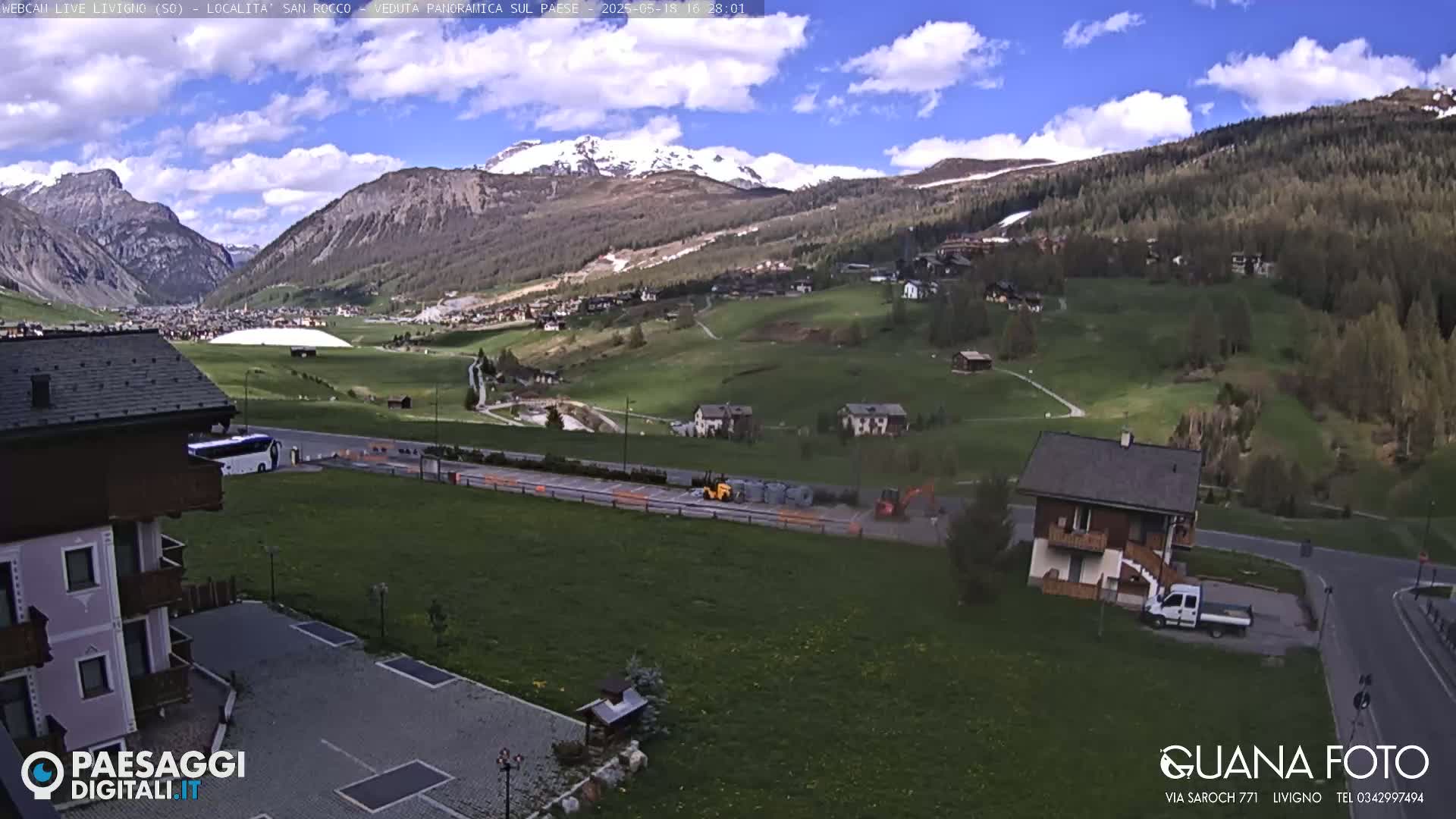 A partly cloudy day reveals a mountain valley town with green fields, a few buildings, and some construction equipment; snow-capped mountains are visible in the background.