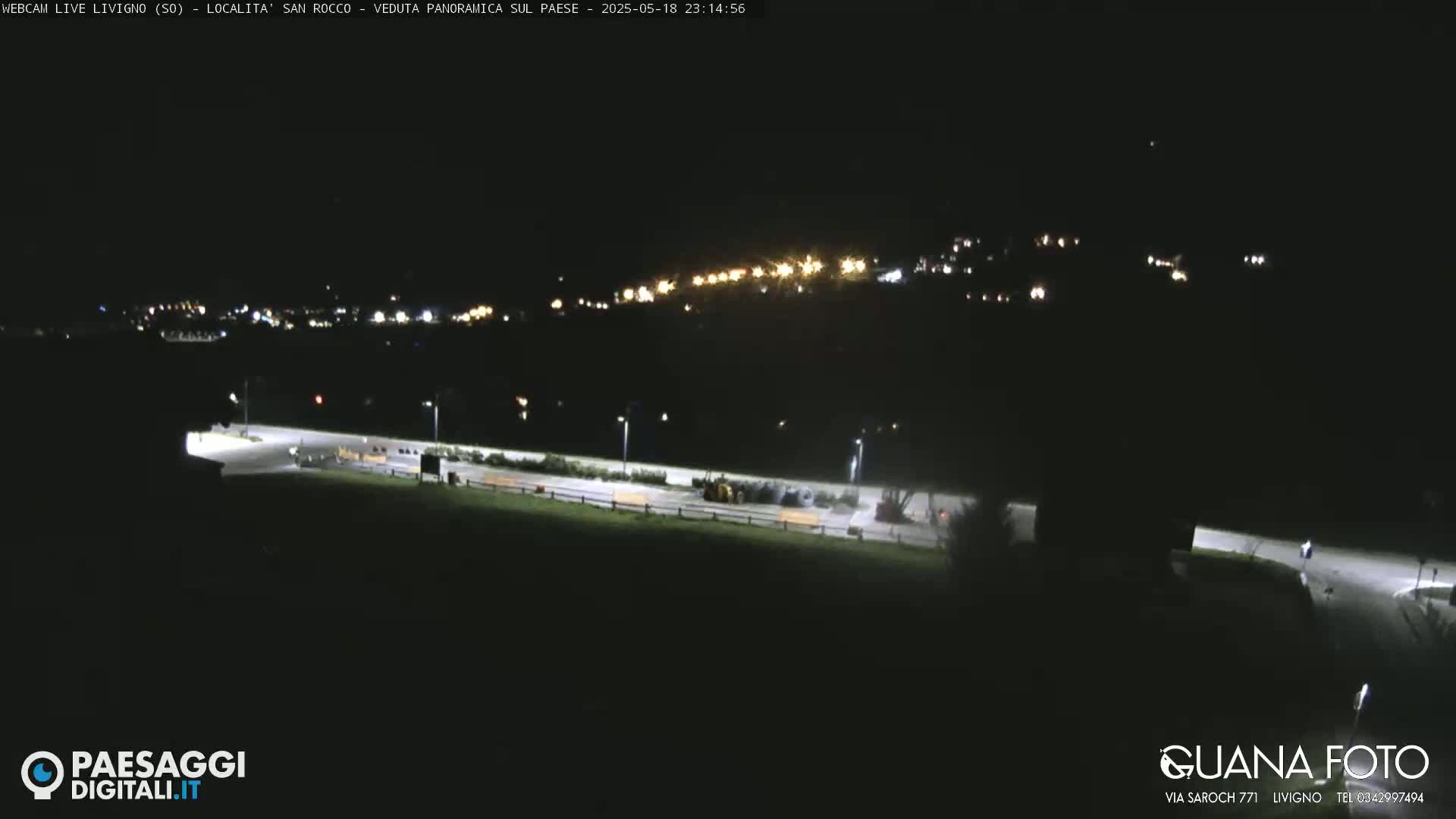A nighttime, long shot of a dimly lit town nestled in a valley, with a few vehicles visible in a parking lot in the foreground.