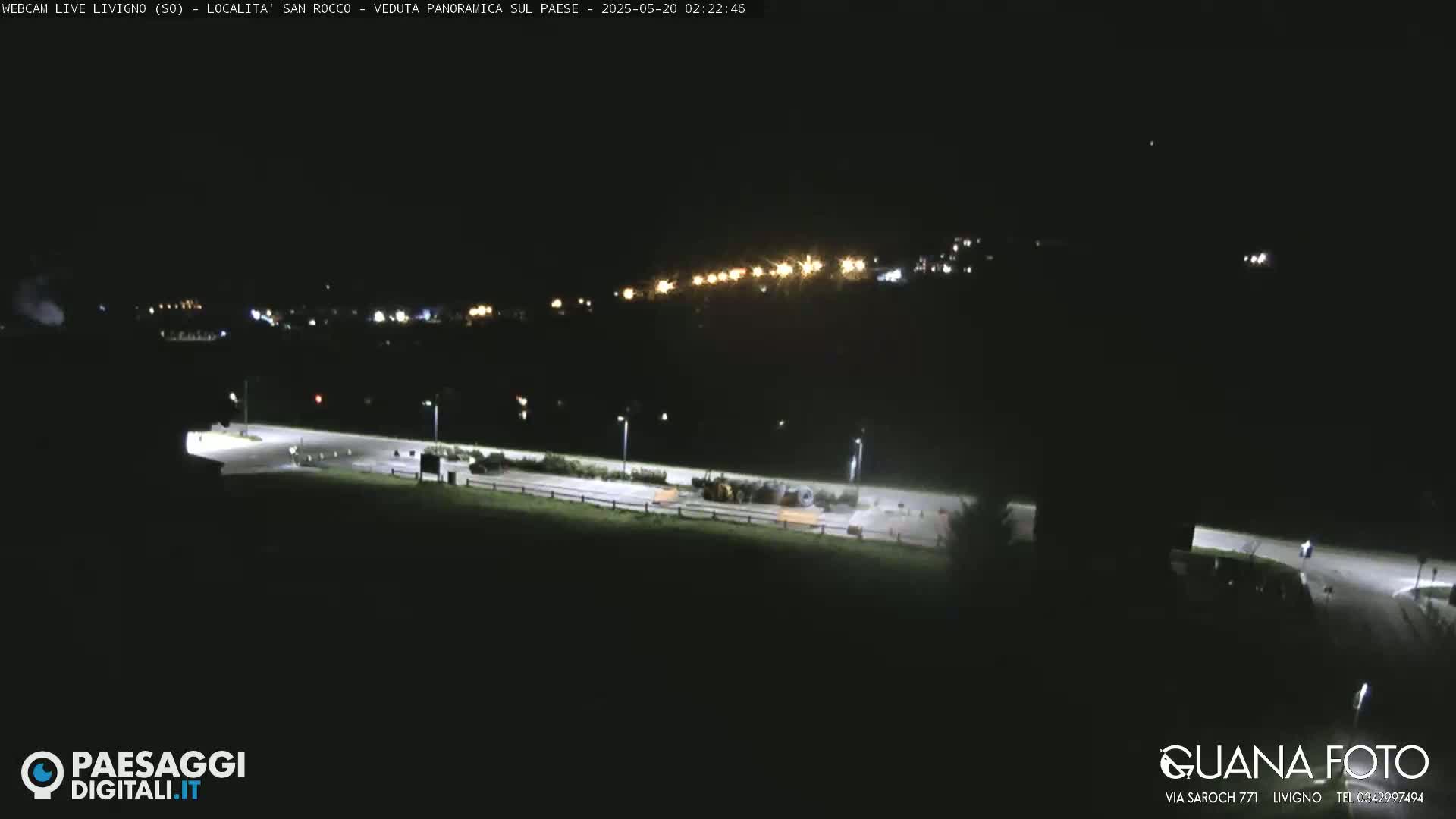 A nighttime, long shot of a mostly dark, empty parking lot and road, with a dimly lit town visible in the distance.