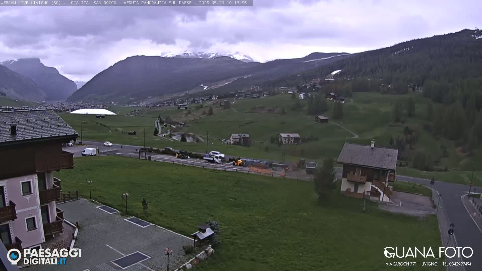 A panoramic view of a village nestled in a valley, surrounded by green hills and snow-capped mountains under an overcast sky.