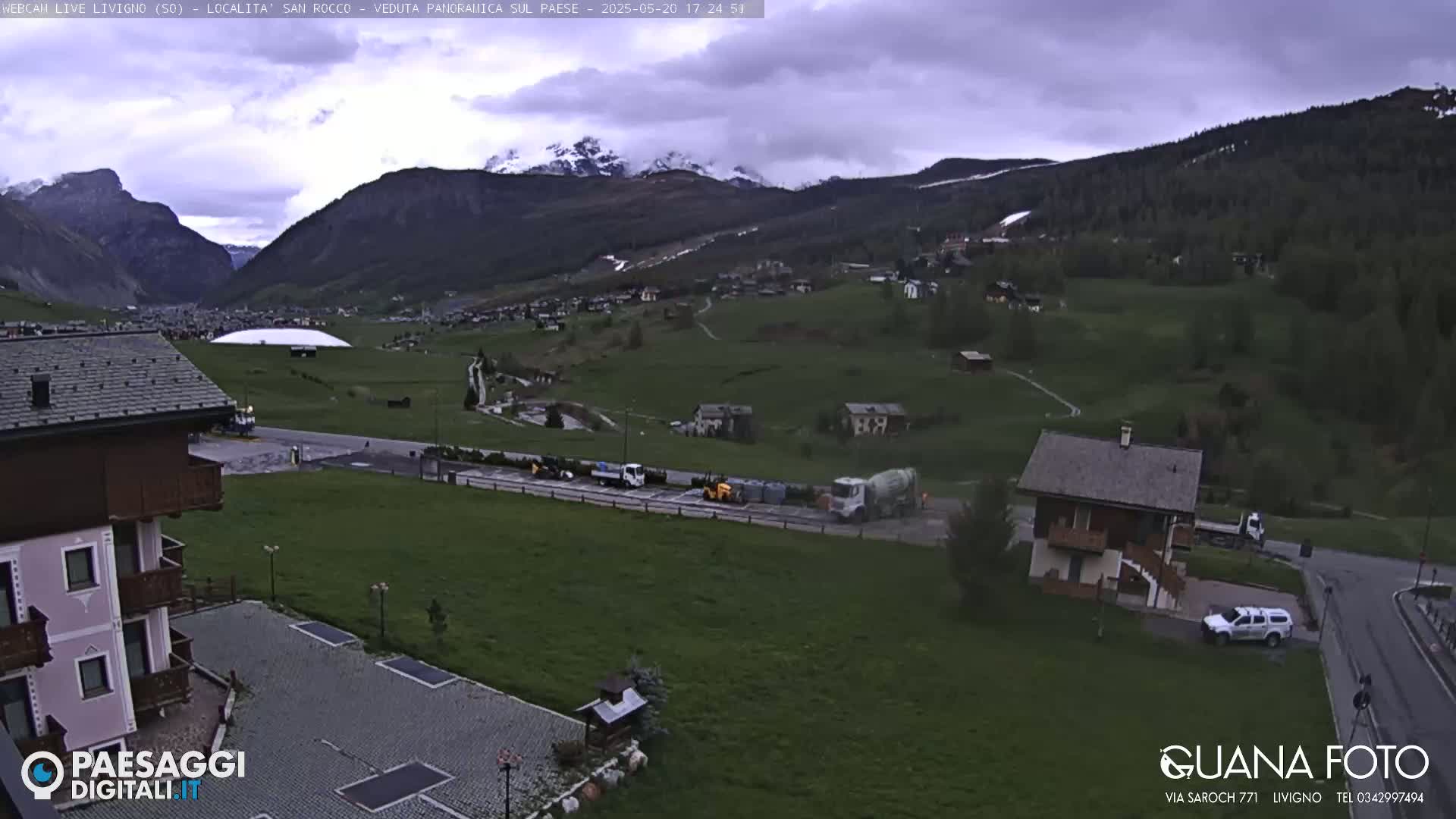 A mountain valley town is shown under an overcast sky, with several vehicles parked near a building and a larger structure in the distance.