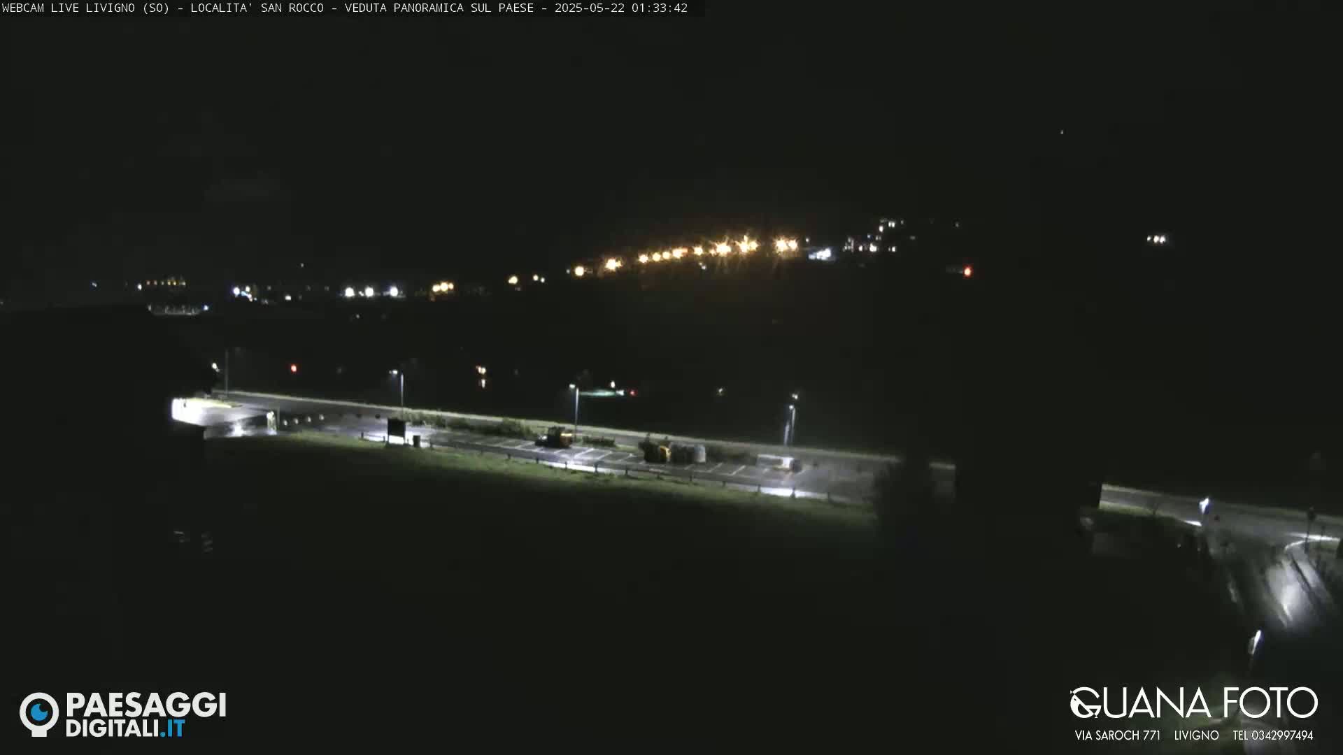 A nighttime long shot of a mostly empty parking lot and road, with a distant town illuminated by lights.