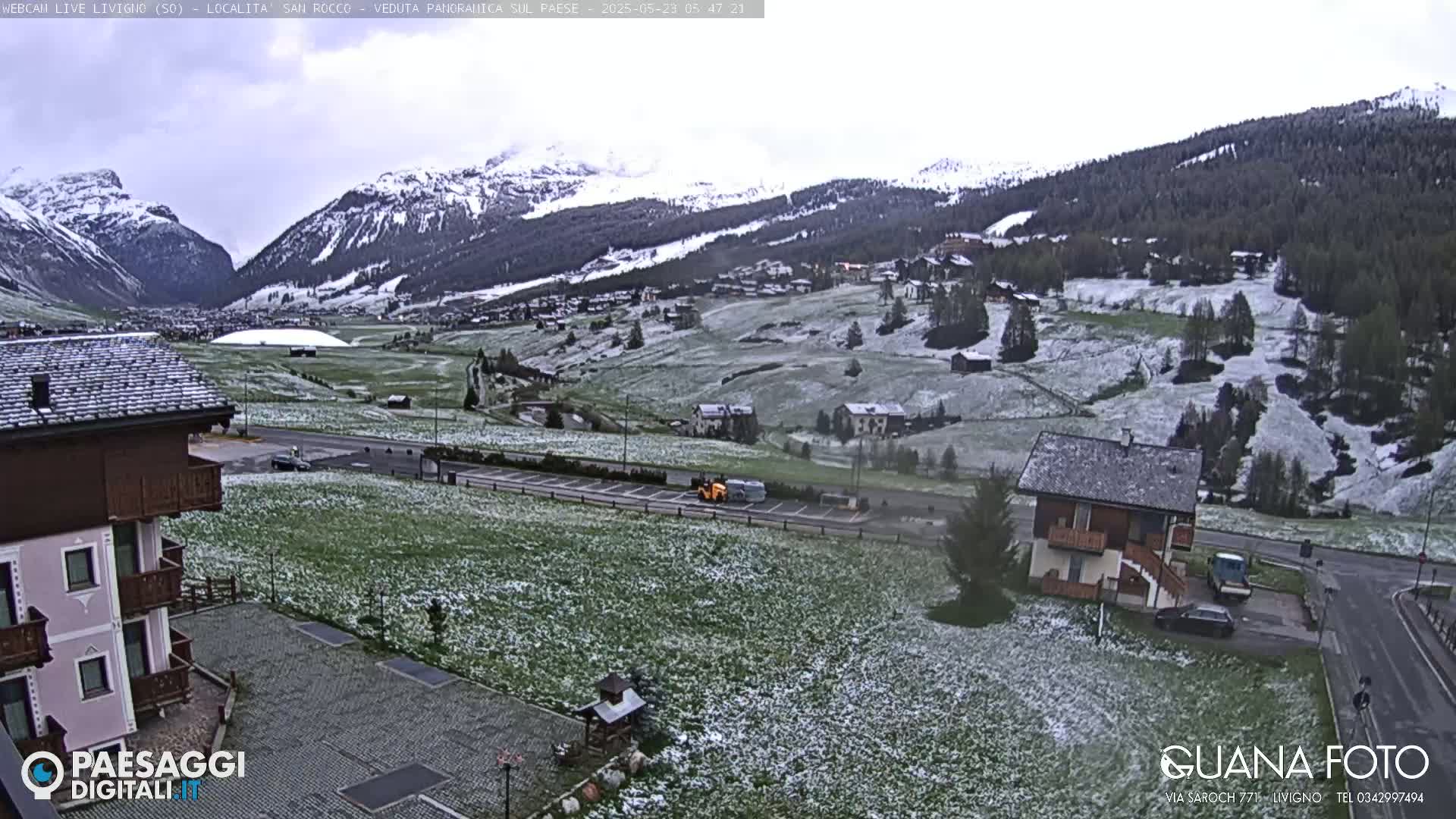 A snow-dusted mountain valley village scene, with green fields speckled with light snow, under an overcast sky.