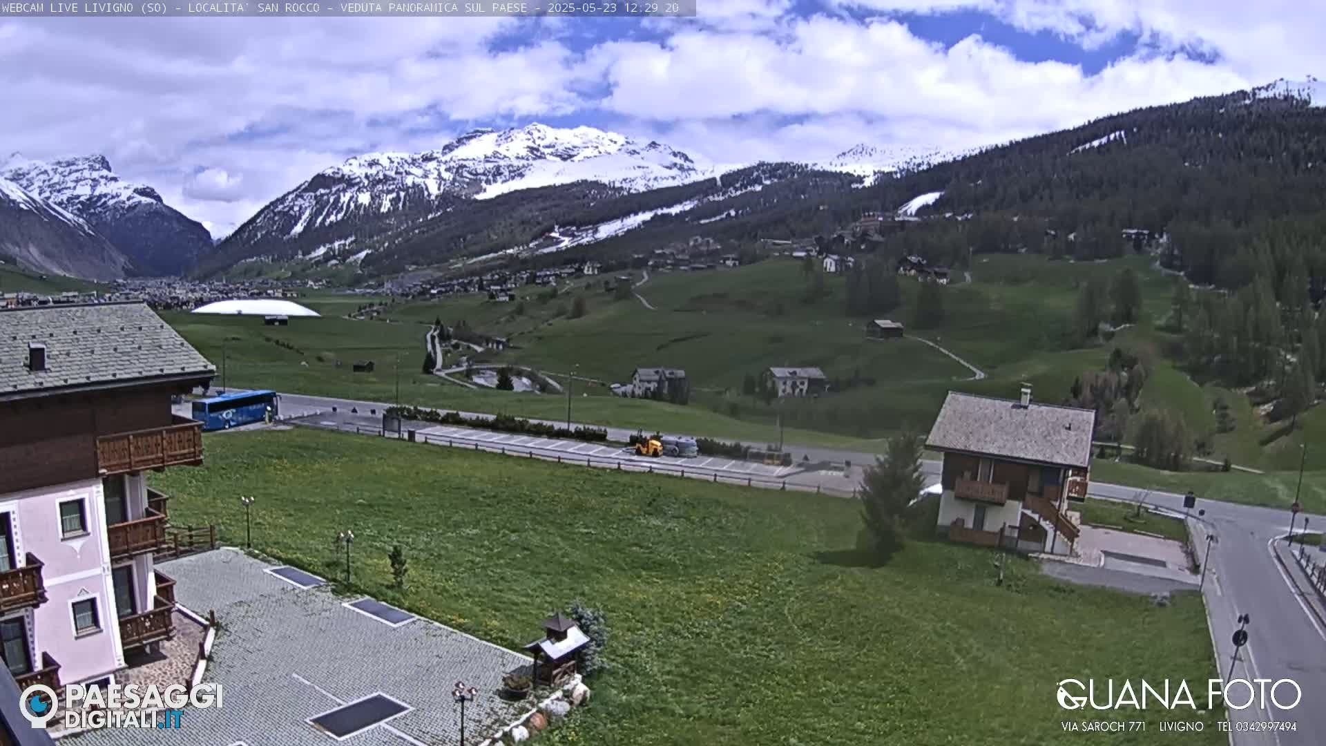 A partly cloudy day reveals a mountain valley with snow-capped peaks, green fields, several buildings, and a blue bus parked near a parking lot.