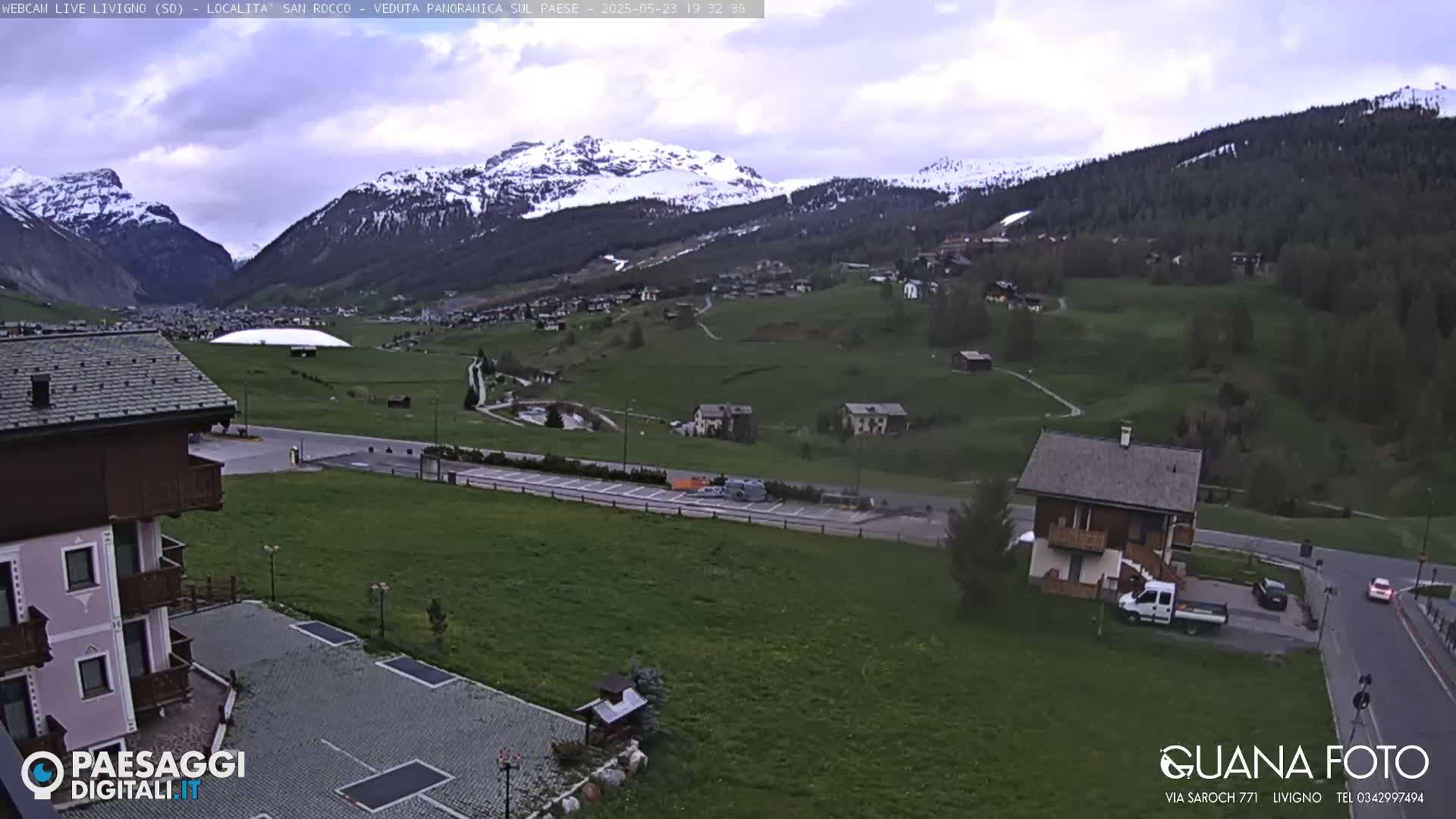 A panoramic view of a village nestled in a valley between snow-capped mountains under an overcast sky.