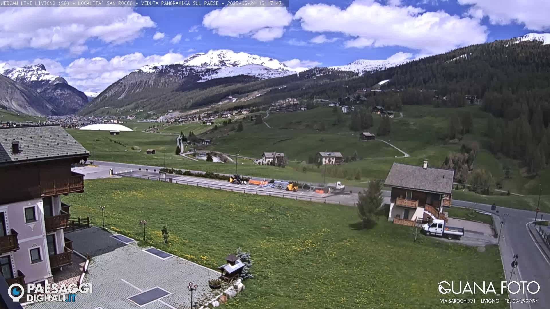 A sunny day in a mountainous region shows a valley with green fields, a few buildings, and snow-capped mountains in the background.