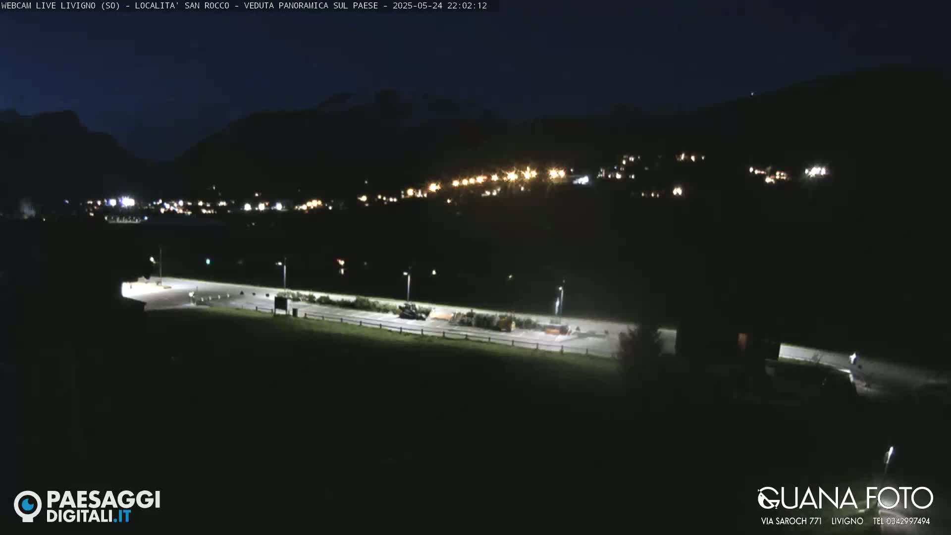 A nighttime view of a village nestled in a valley between mountains, illuminated by scattered lights, under a clear, dark sky.