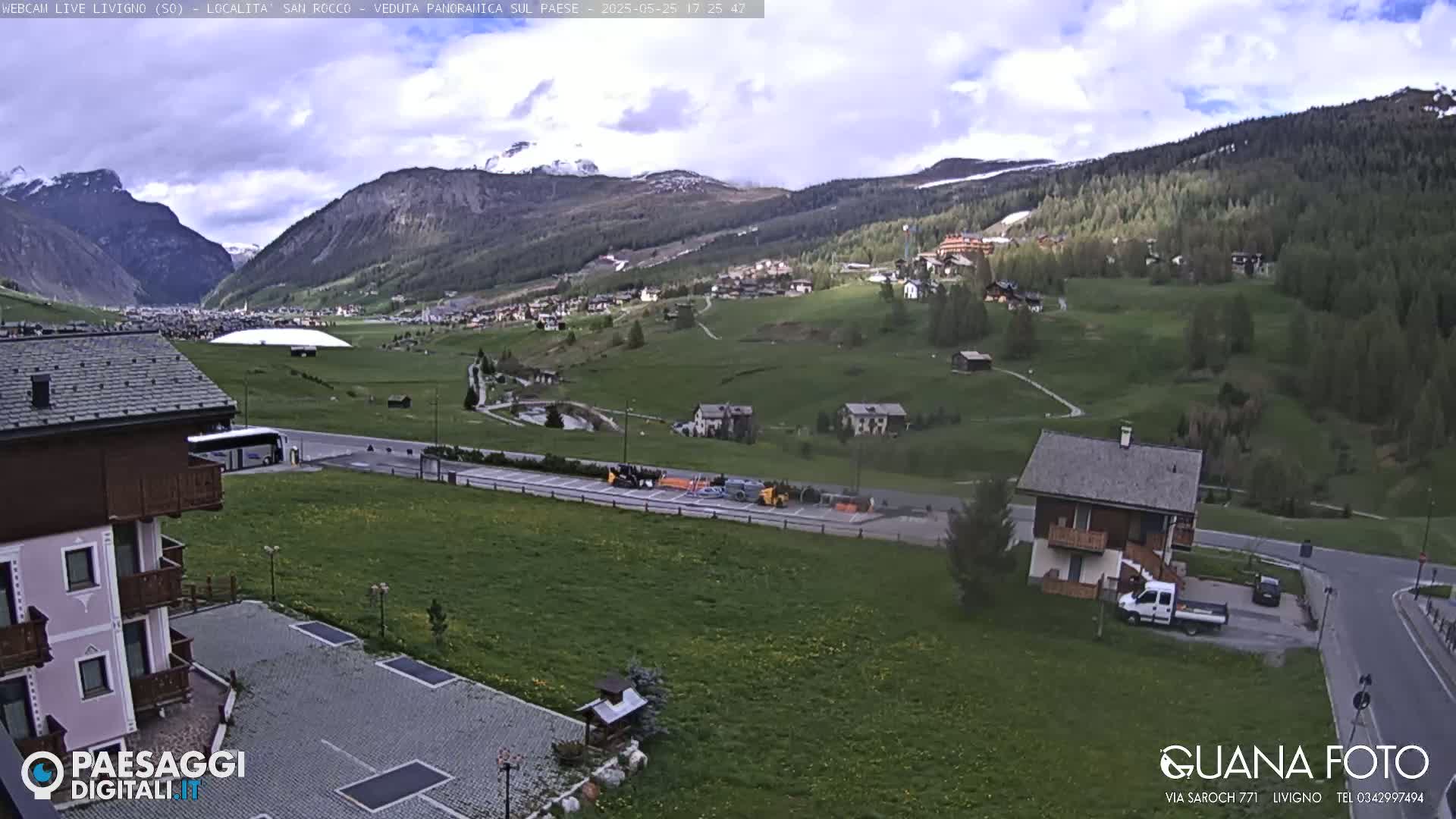 A partly cloudy day reveals a valley town nestled between snow-capped mountains, with a large building, vehicles, and a few houses visible.