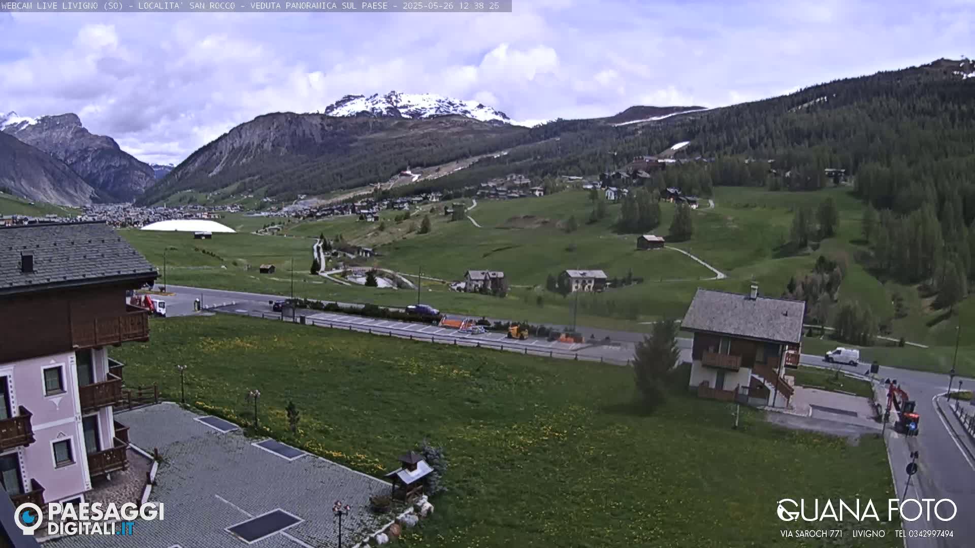 A partly cloudy day reveals a valley town nestled among green hills and snow-capped mountains.