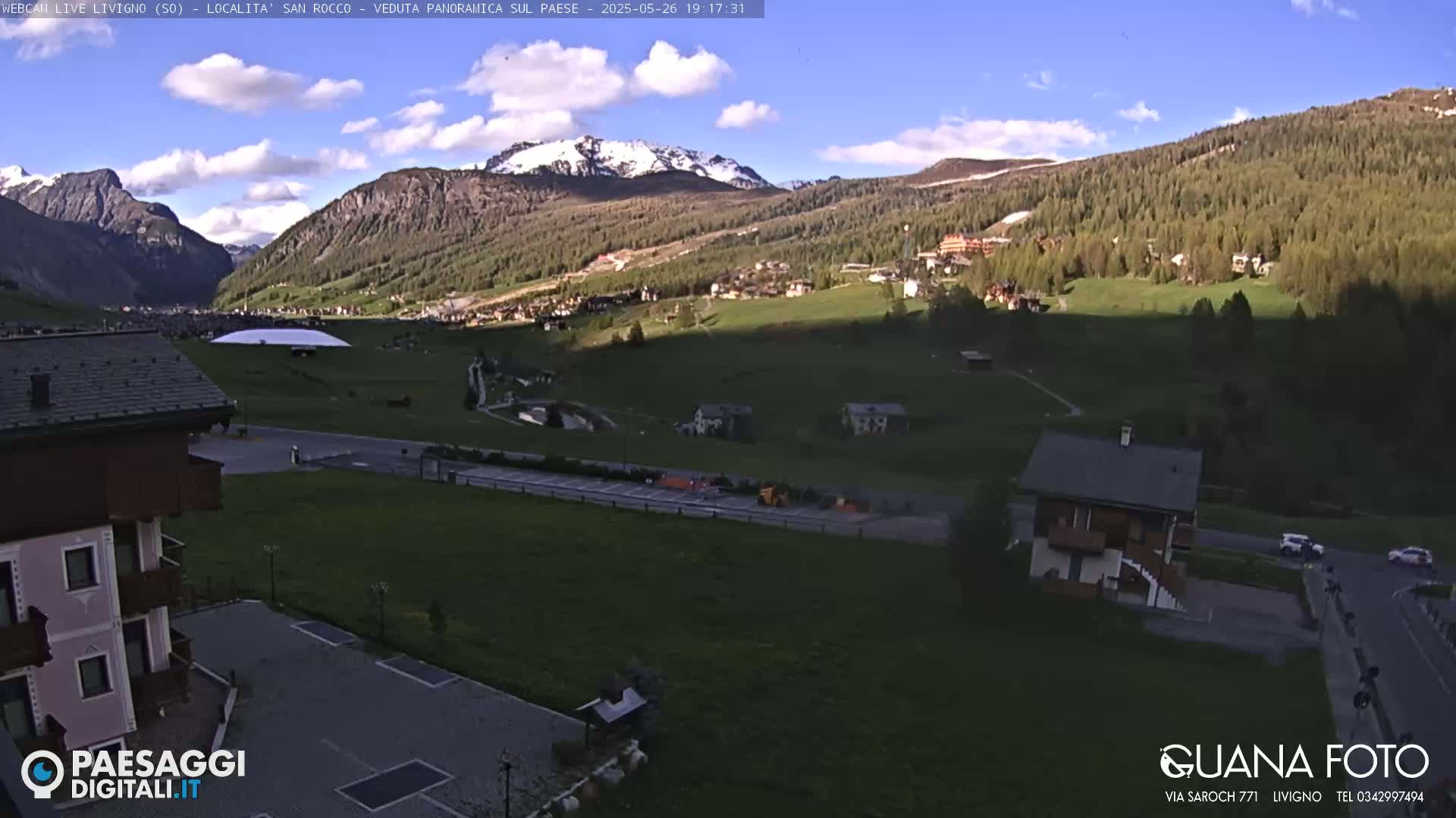 A sunny, partly cloudy view of a mountain valley town with snow-capped mountains in the background.