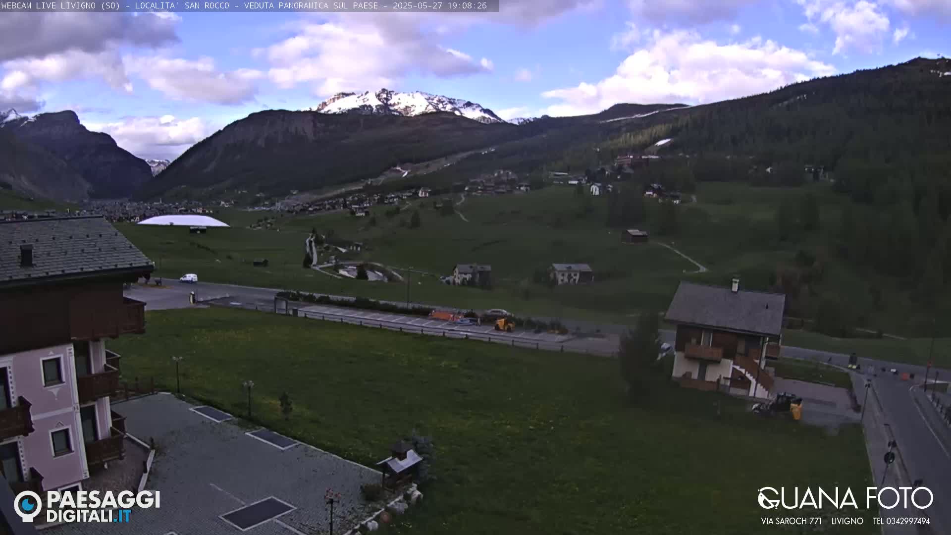 A partly cloudy daytime view of a village nestled in a valley between snow-capped mountains.