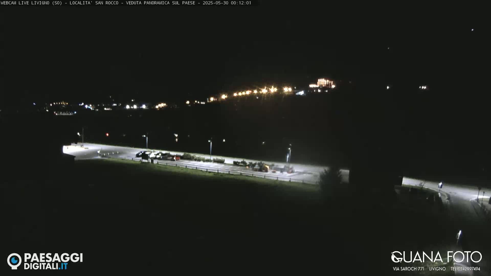A nighttime, long shot of a parking lot with some vehicles, illuminated by artificial lights, overlooking a hillside with distant building lights.