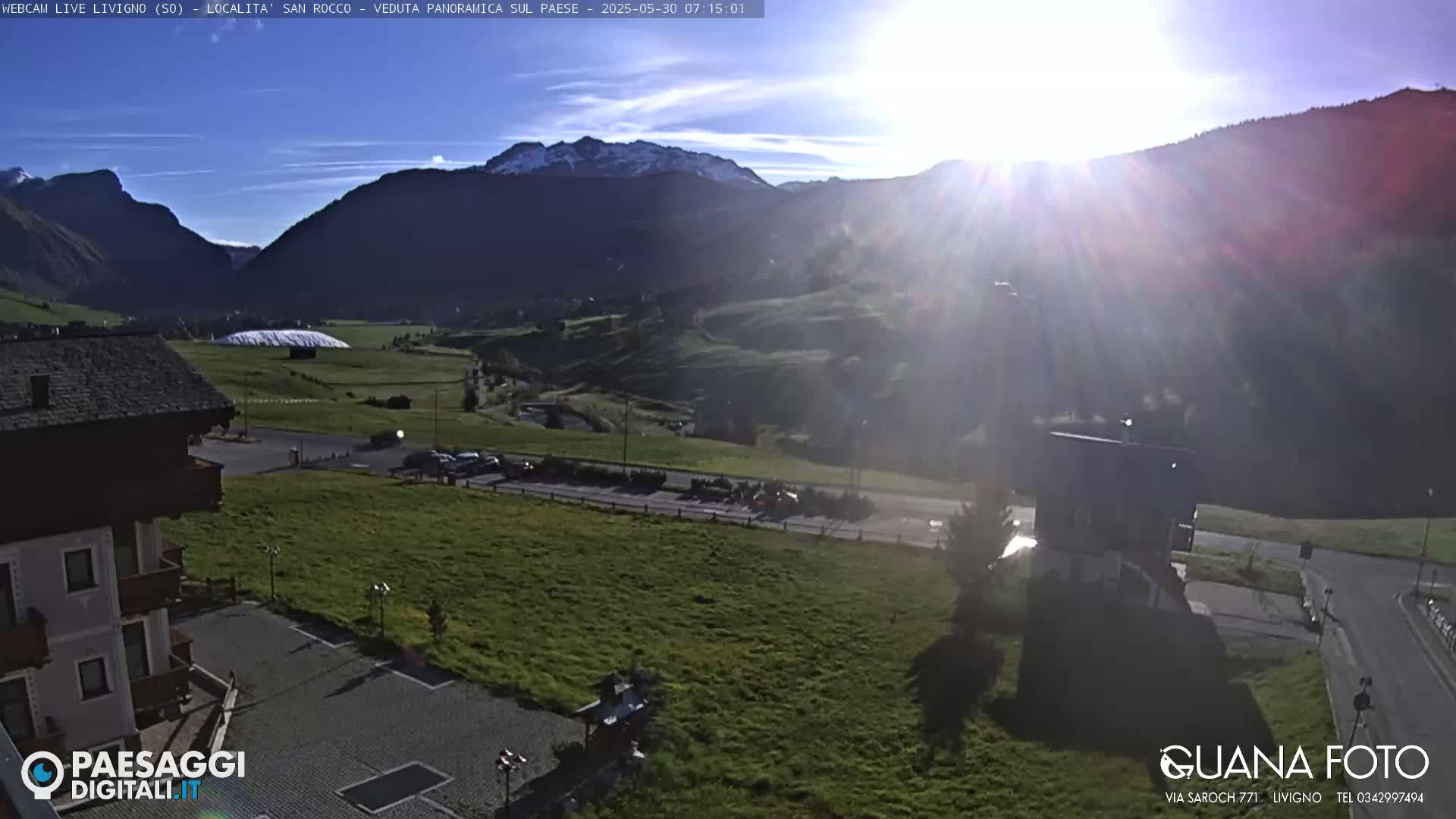A sunny, mountain vista with a partially snow-capped mountain range in the background, overlooking a green valley with buildings, parking lots, and a road.