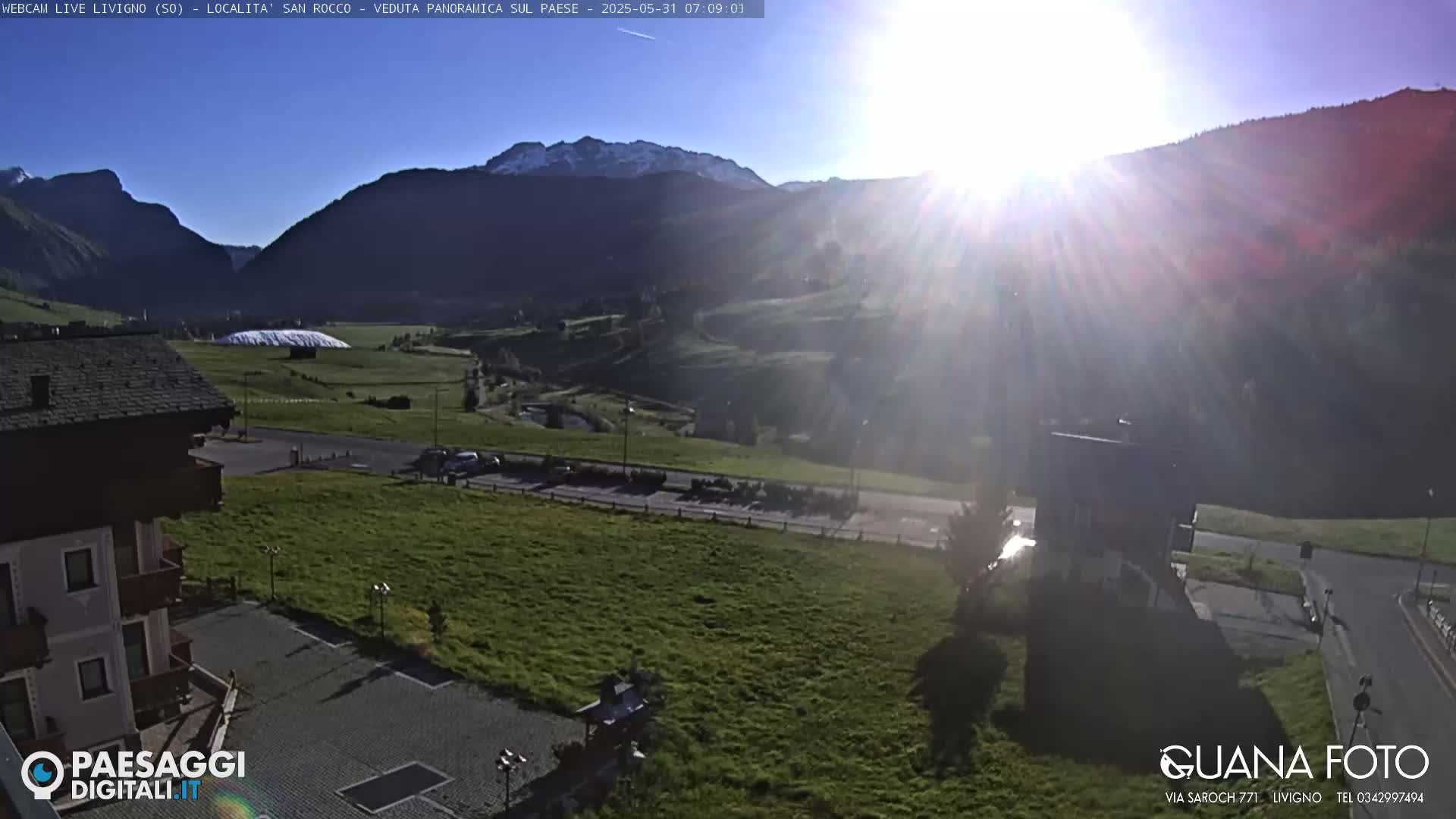 A sunny, bright morning view overlooking a green valley and mountains in the distance, with a few buildings and a road visible in the foreground.
