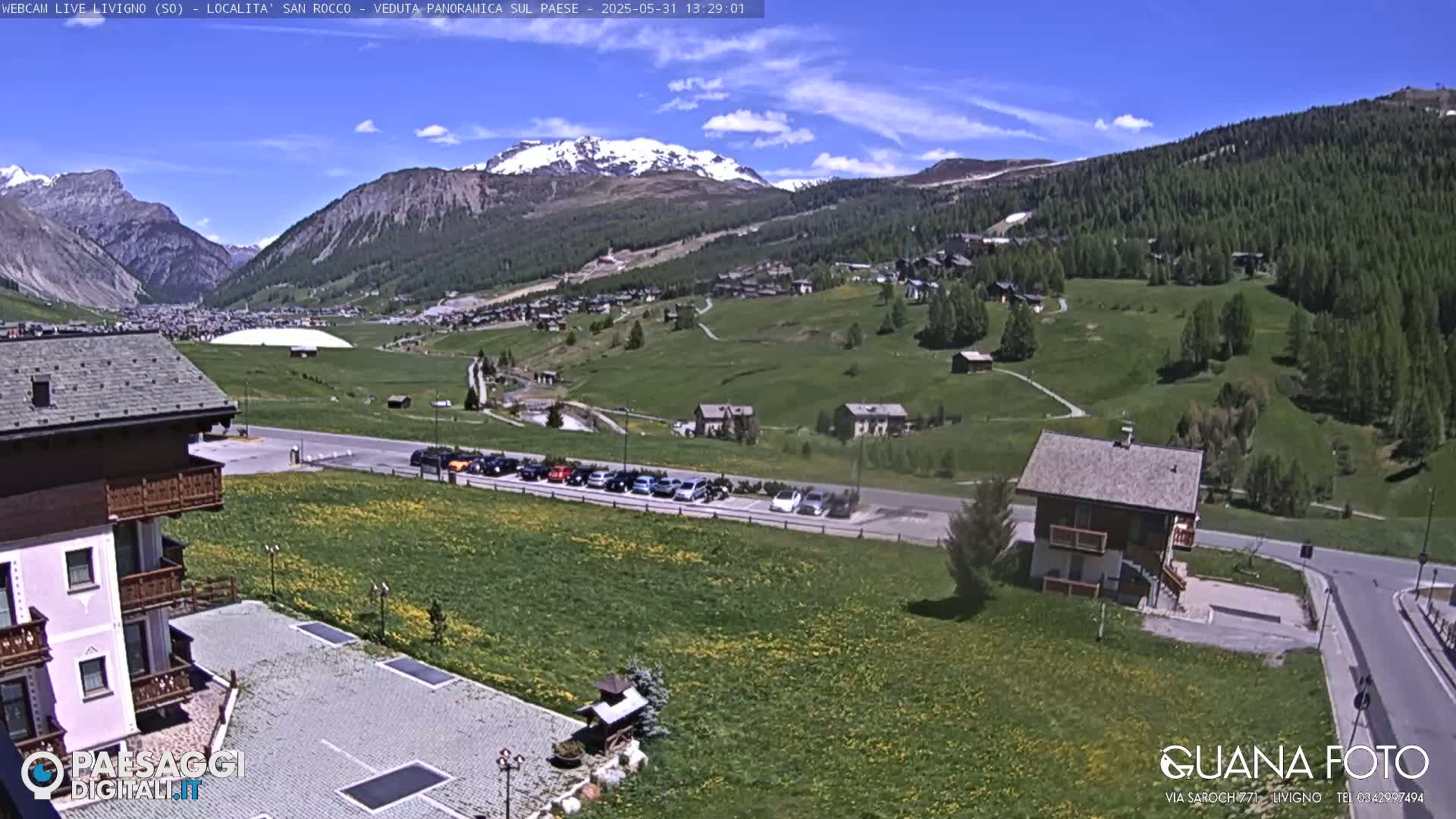 A sunny mountain valley town is viewed from above, showing a parking lot, buildings, and green fields, with snow-capped mountains in the background.